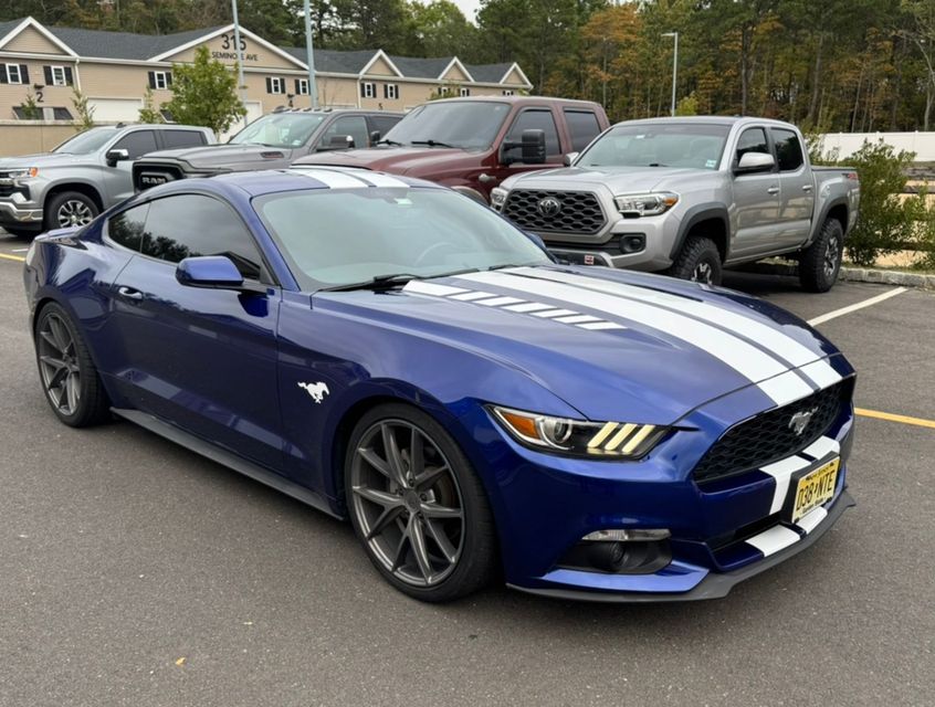 Blue Ford Mustang parked in a lot, with silver pickup trucks and houses in the background
