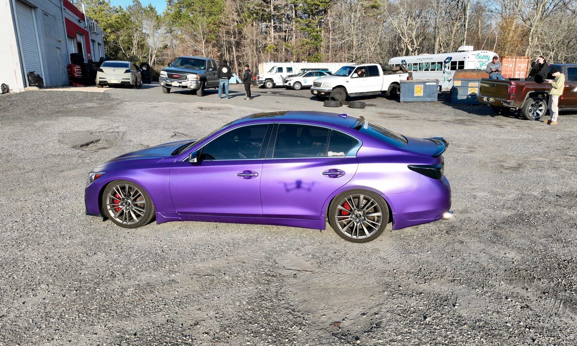 Purple sports sedan parked on gravel lot with trucks and buildings in the background