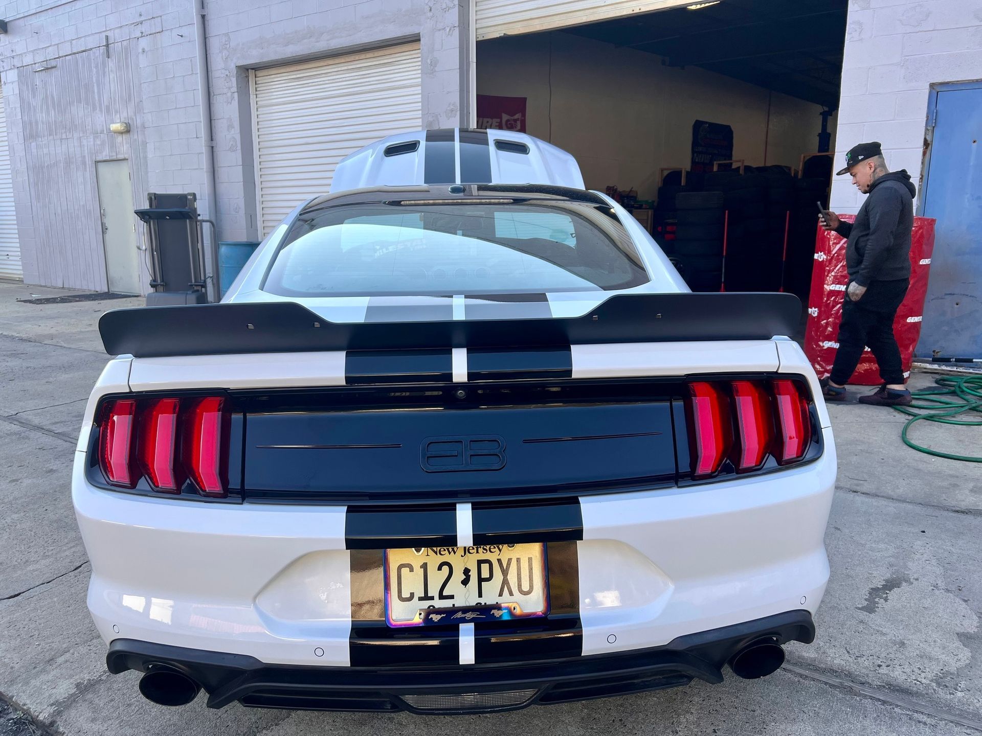 White Ford Mustang rear view with black racing stripes, parked outside a garage with a person nearby