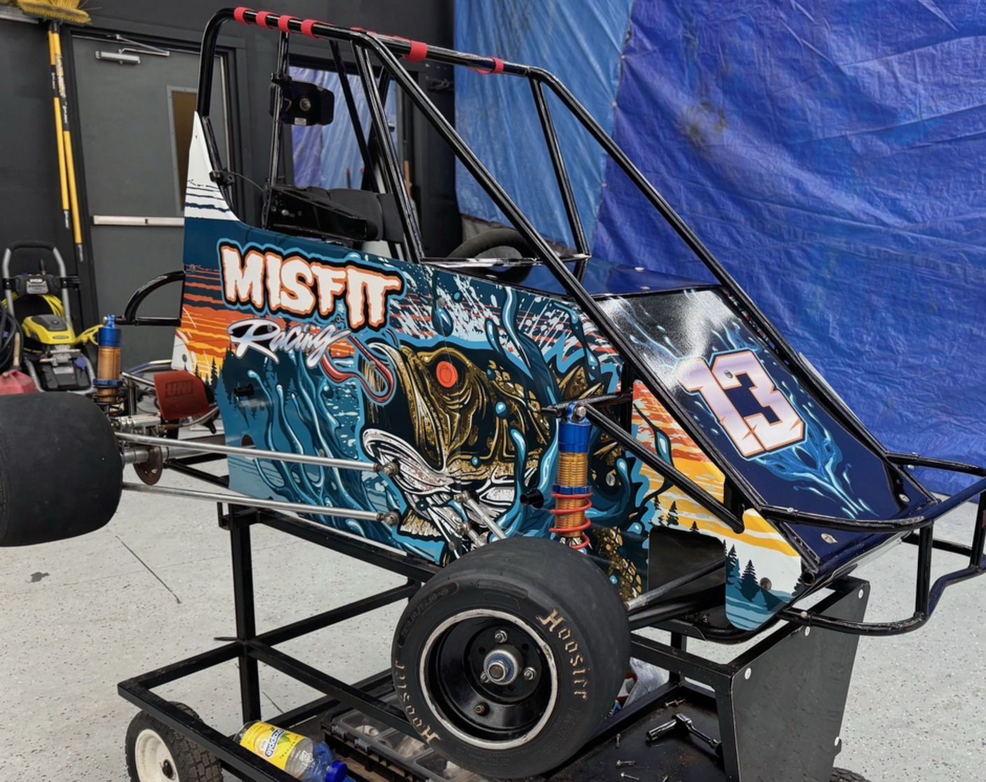 Blue MISFIT race car chassis with large front wheel in a garage, featuring a colorful graphic wrap