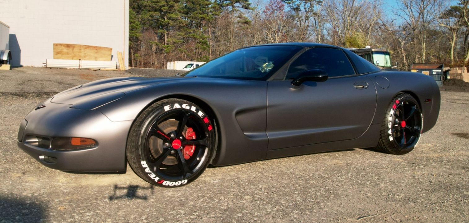 Gray sports car with black-and-red rims parked on a gravel lot in sunlight