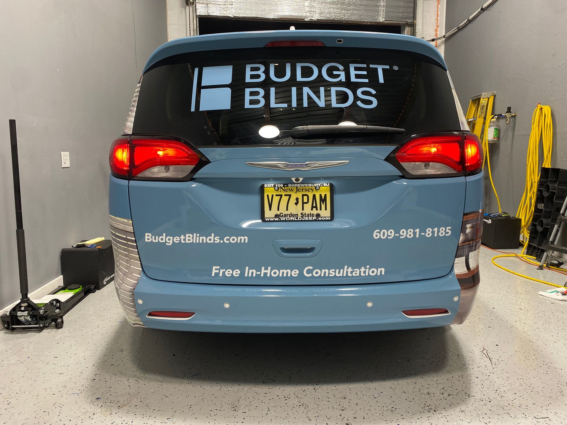 Blue Budget Blinds van parked indoors, rear view with company logo and license plate.