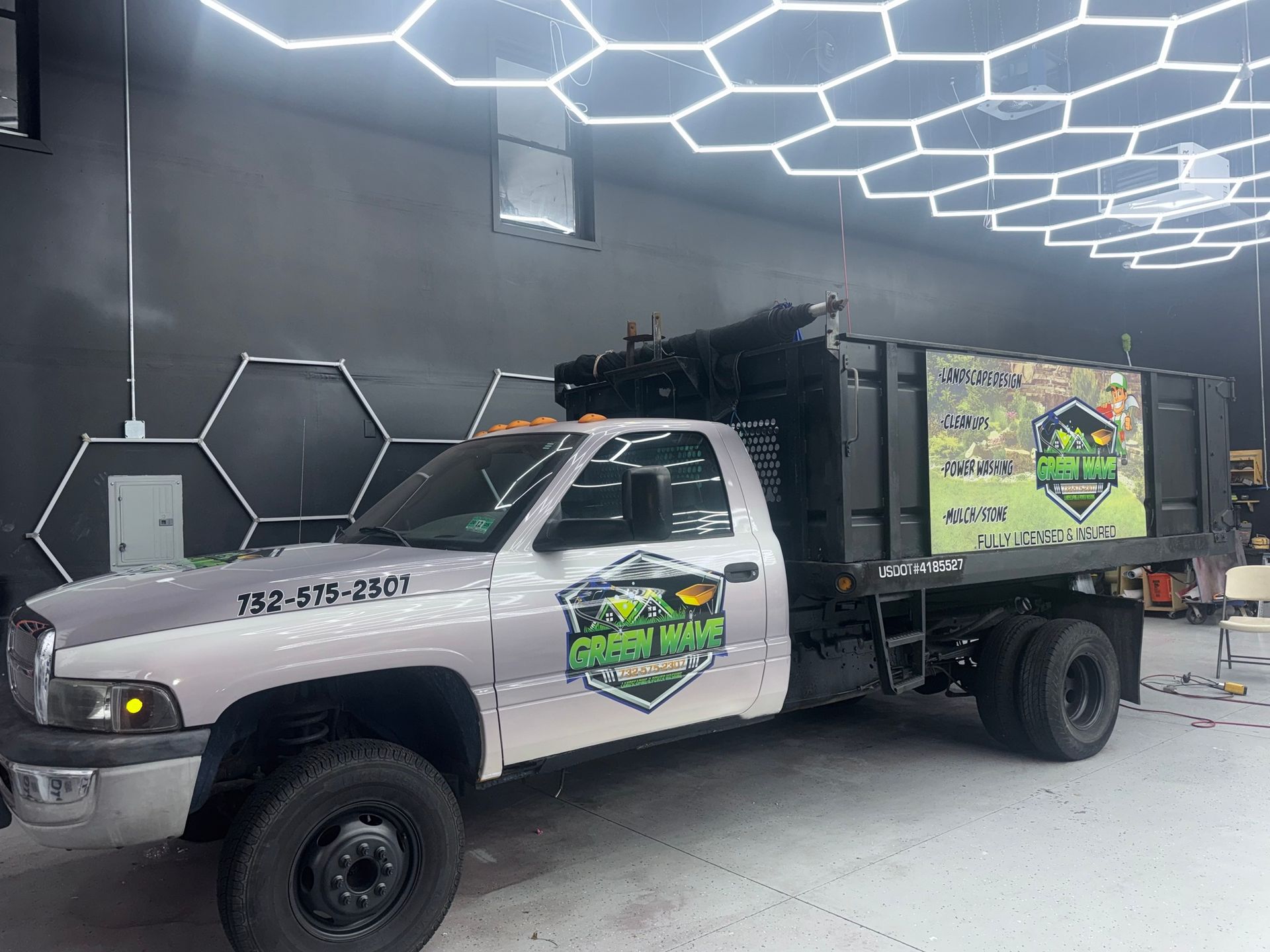 White utility truck with a black dump bed and green logo parked indoors under hexagon ceiling lights