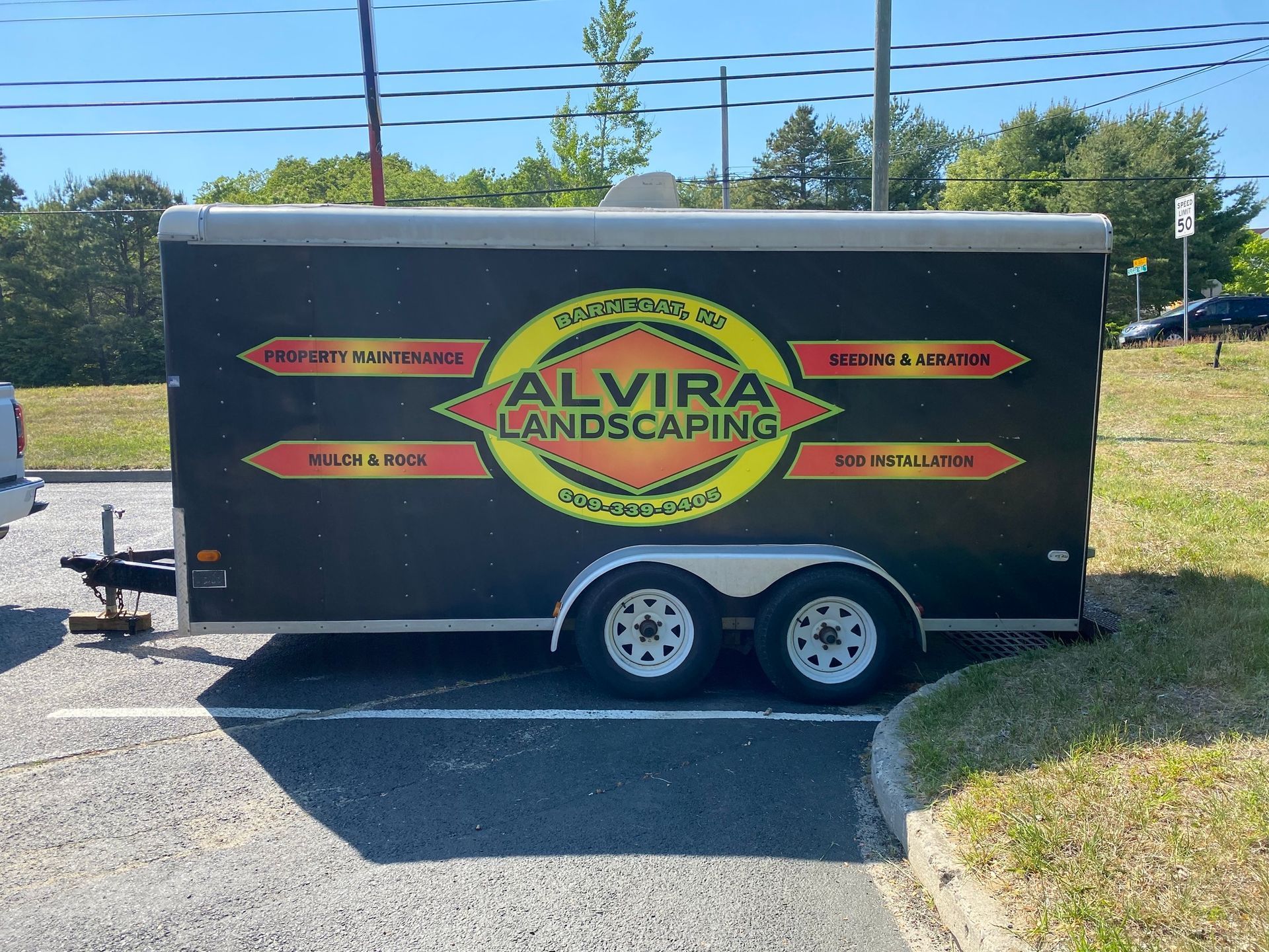 Black landscaping trailer with “Alvira Landscaping” logo parked in a lot, white wheels and trim.