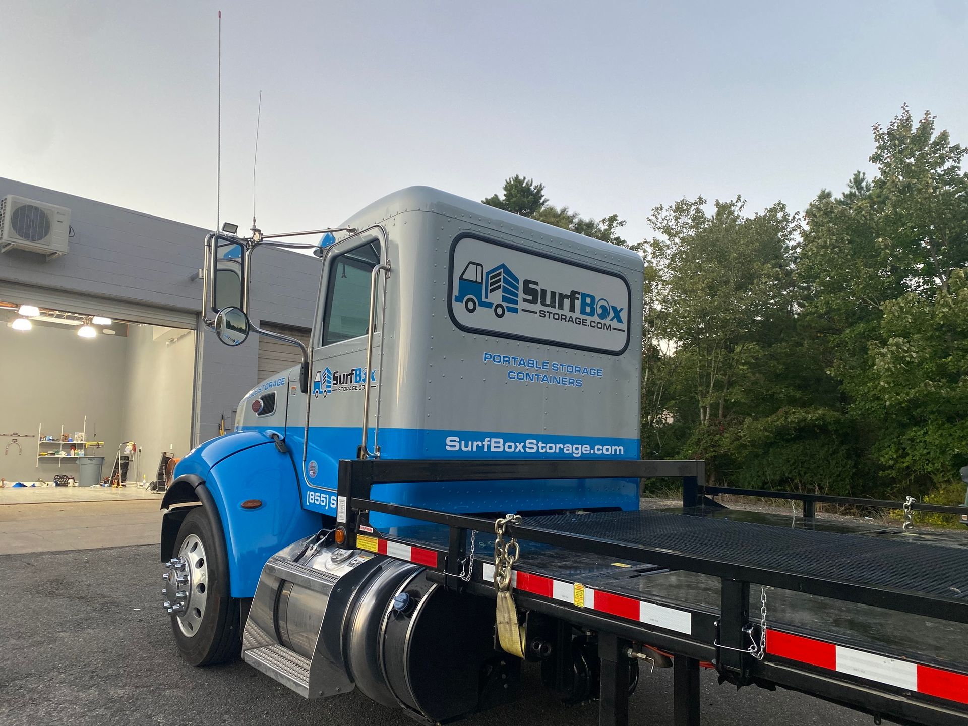 Blue semi-truck with flatbed trailer parked beside a building at dusk, viewed from the rear side.