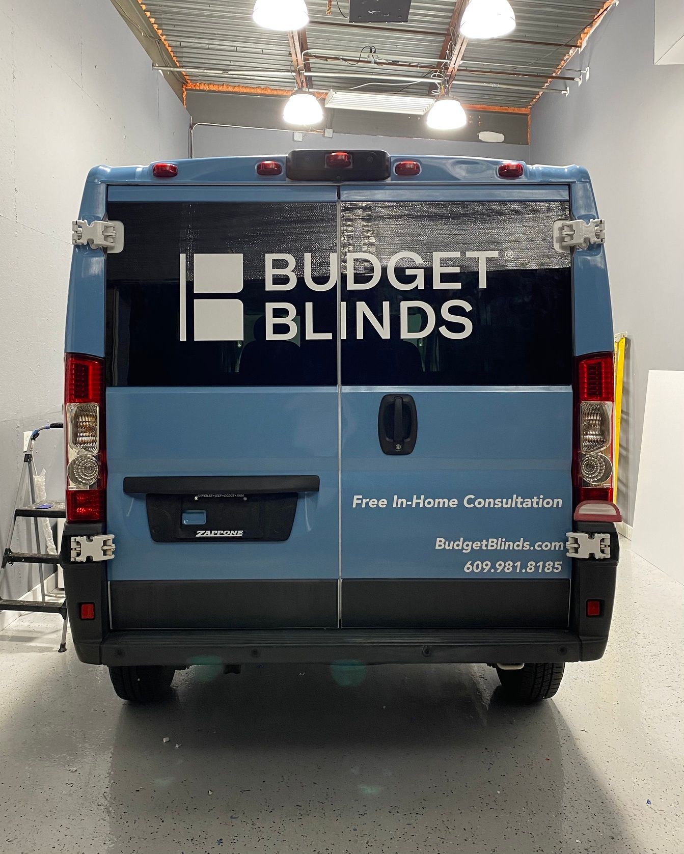 Budget Blinds delivery van, dark blue and light blue, parked inside a garage bay.