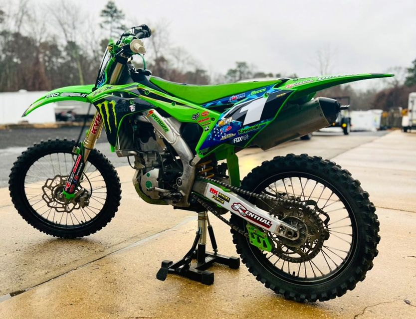 Green dirt bike on a stand outdoors, viewed from the side on wet pavement.