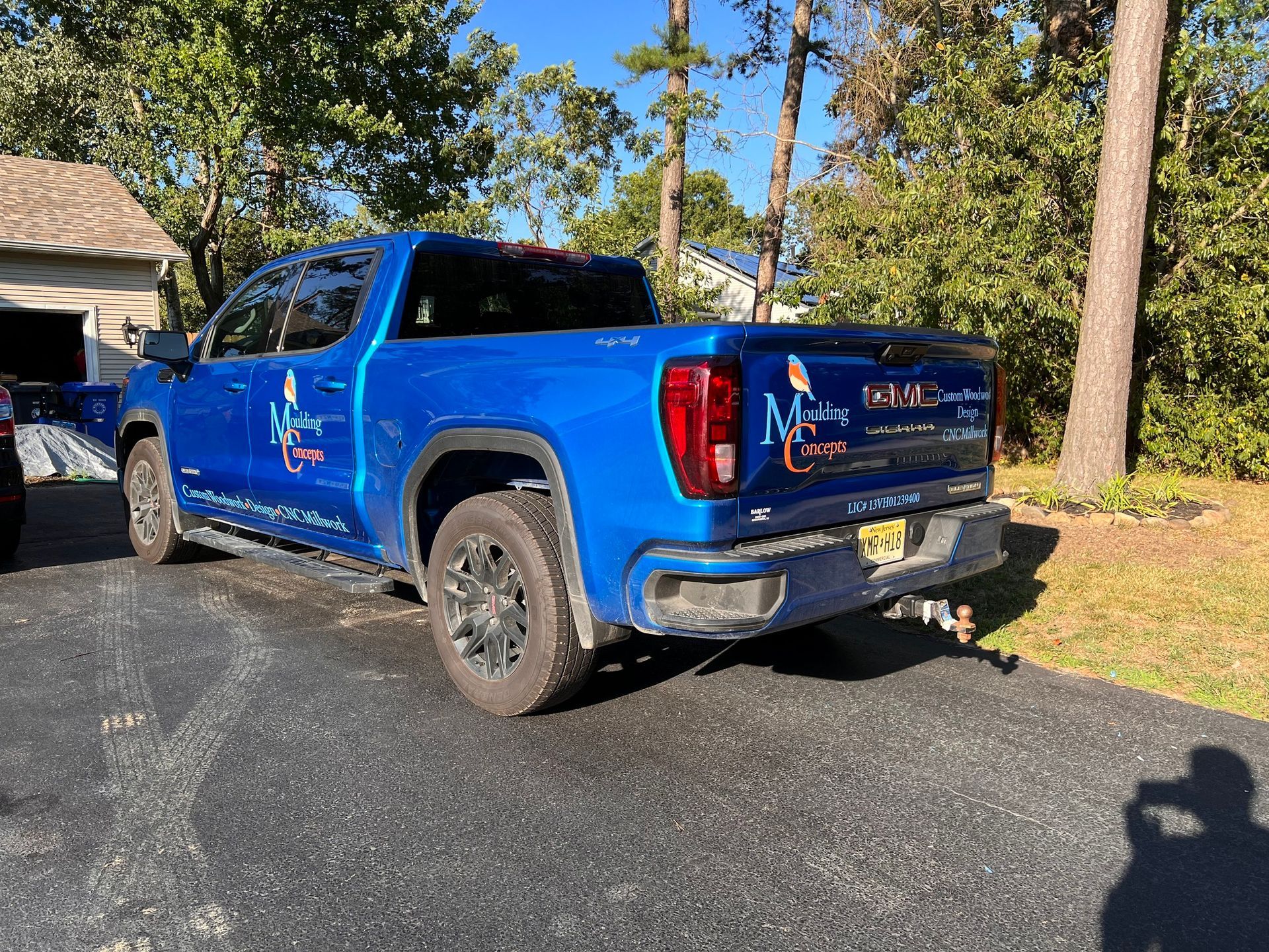 Blue pickup truck parked on a driveway beside trees and a house, with sunlight casting shadows.