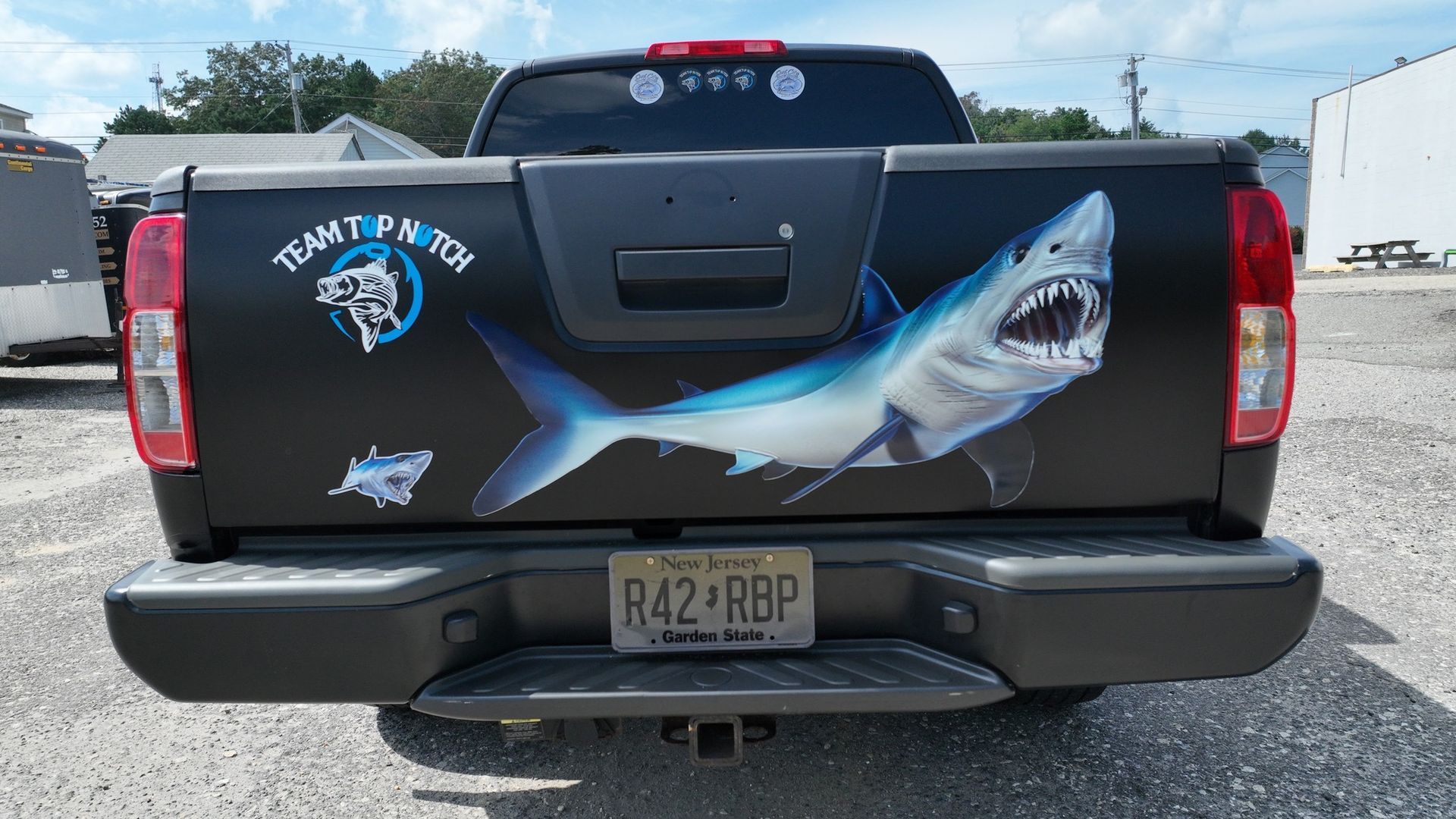 Black pickup truck tailgate with large shark mural and logo, parked outdoors behind visible license plate.