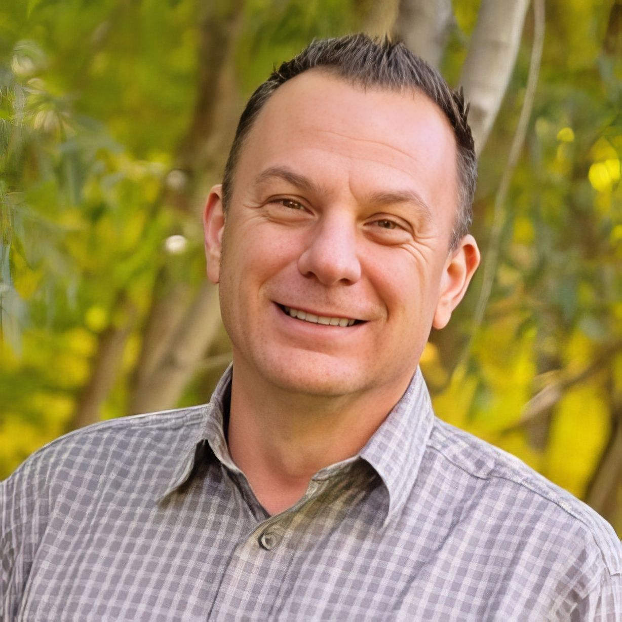 Man smiling in a gray plaid shirt outdoors with blurred green trees behind him