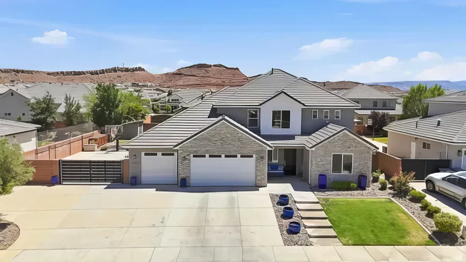 Suburban two-story house with gray roof, white garage, desert hills, and landscaped front yard under clear sky