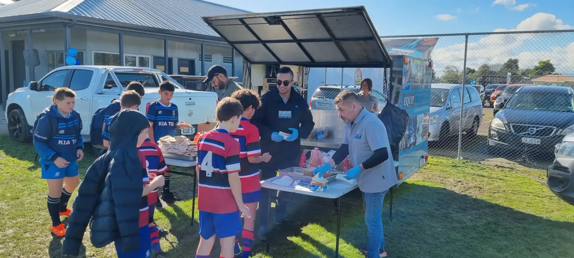 Children in sports uniforms at an outdoor snack table under a canopy on a sunny day