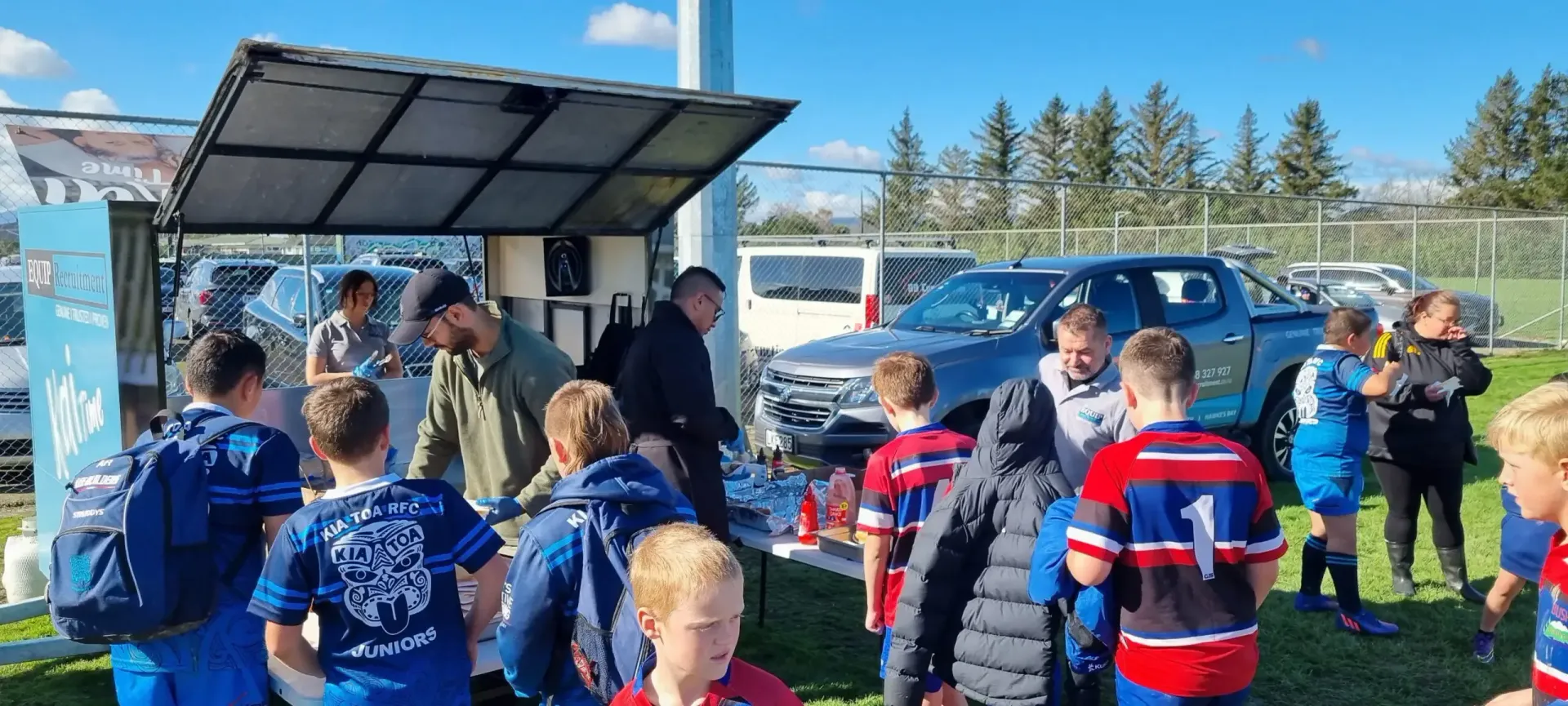 People gathered around a food truck in a sunny outdoor field, some wearing blue sports jerseys.