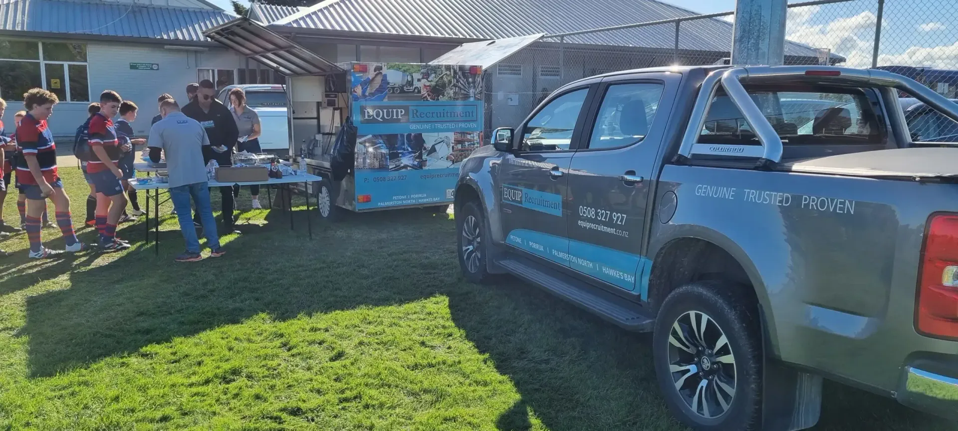 People near a blue pickup truck at an outdoor event booth on a sunny day