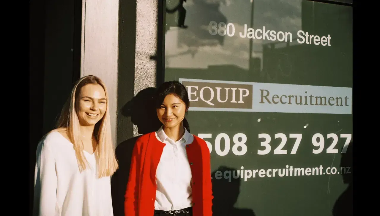 Two smiling people standing outside an Equif Recruitment office with a dark green sign