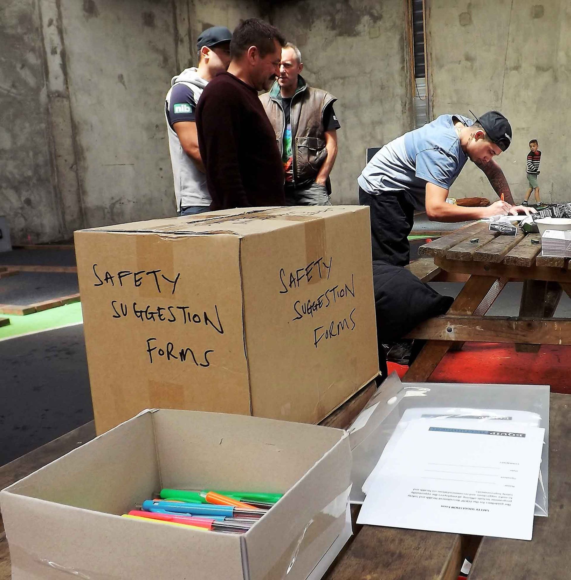 Workers packing safety suggestion forms and pens into boxes at a table in an indoor industrial space