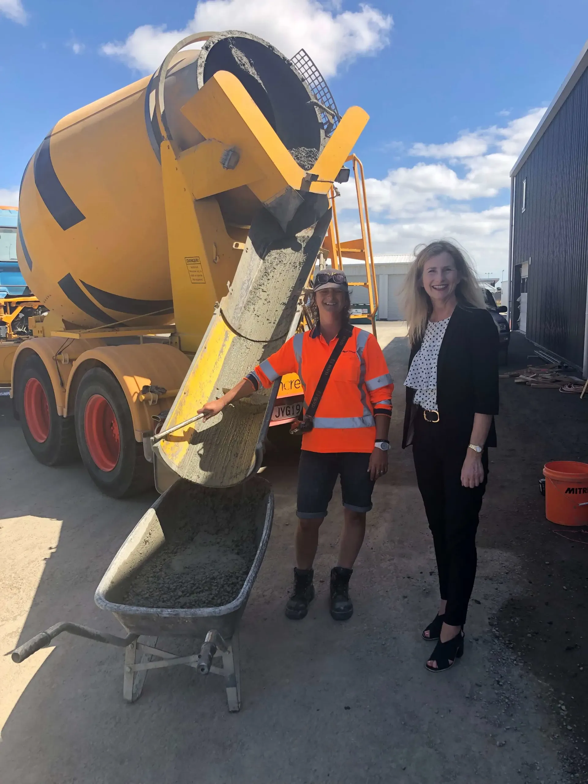 Worker and woman beside a yellow cement mixer pouring concrete into a wheelbarrow outdoors.