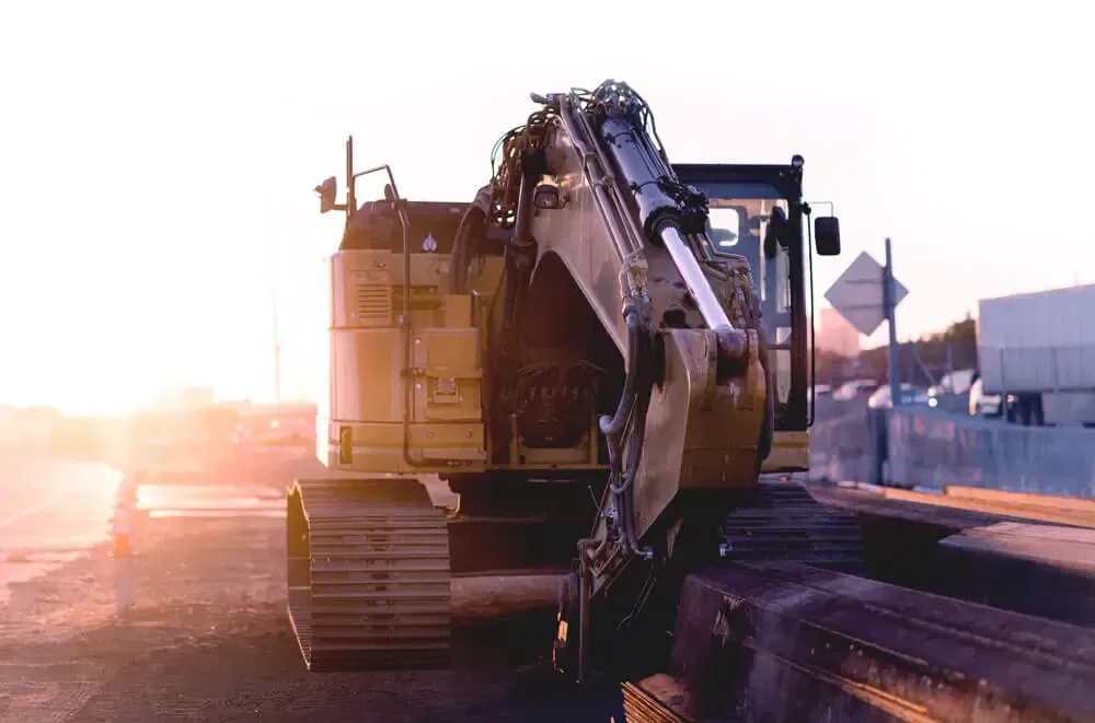 Excavator loading material at a construction site during sunset, with bright glare on the left.