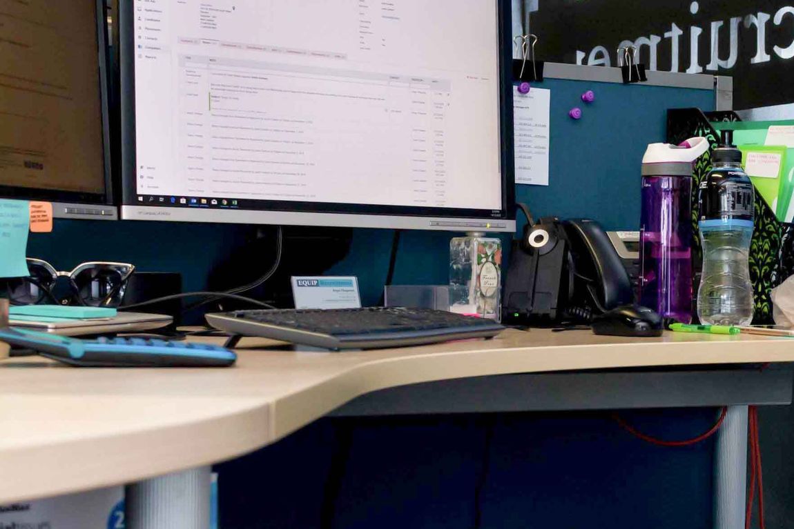 Office desk with dual monitors, keyboard, mouse, and water bottles on a cluttered workspace.