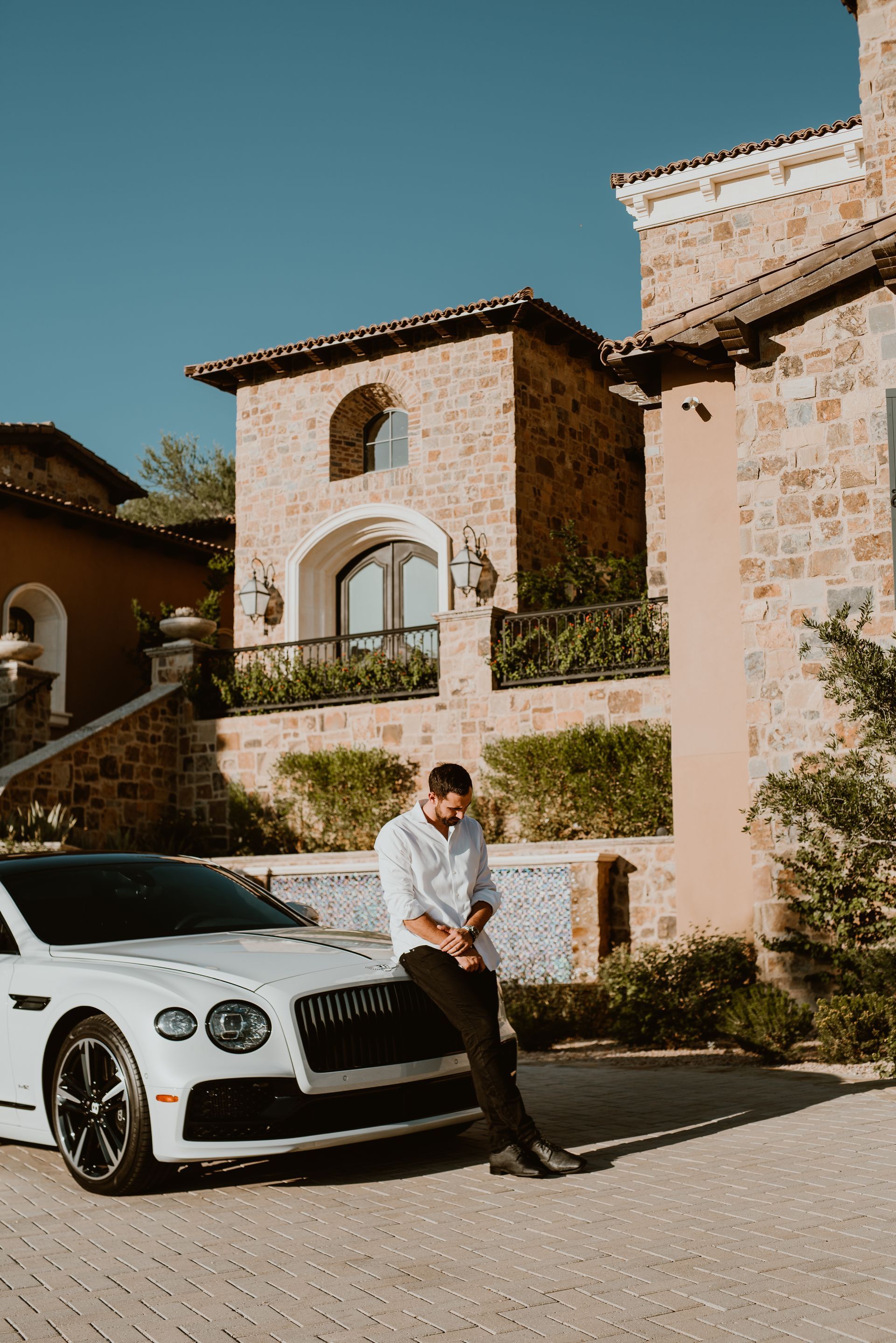 A man is sitting on the hood of a white car in front of a house.