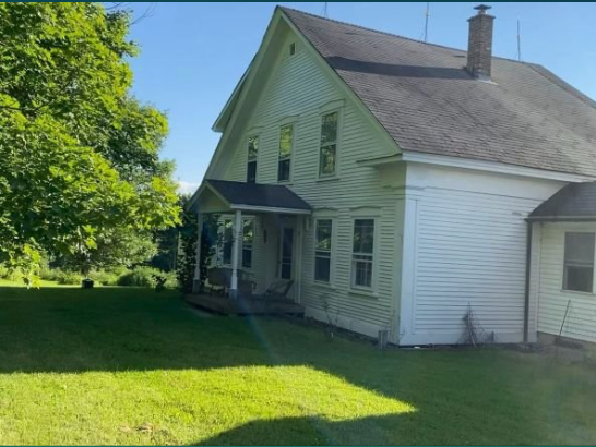 Two-story, white house with a porch, set in a green, grassy yard. A blue sky and trees are in the background.