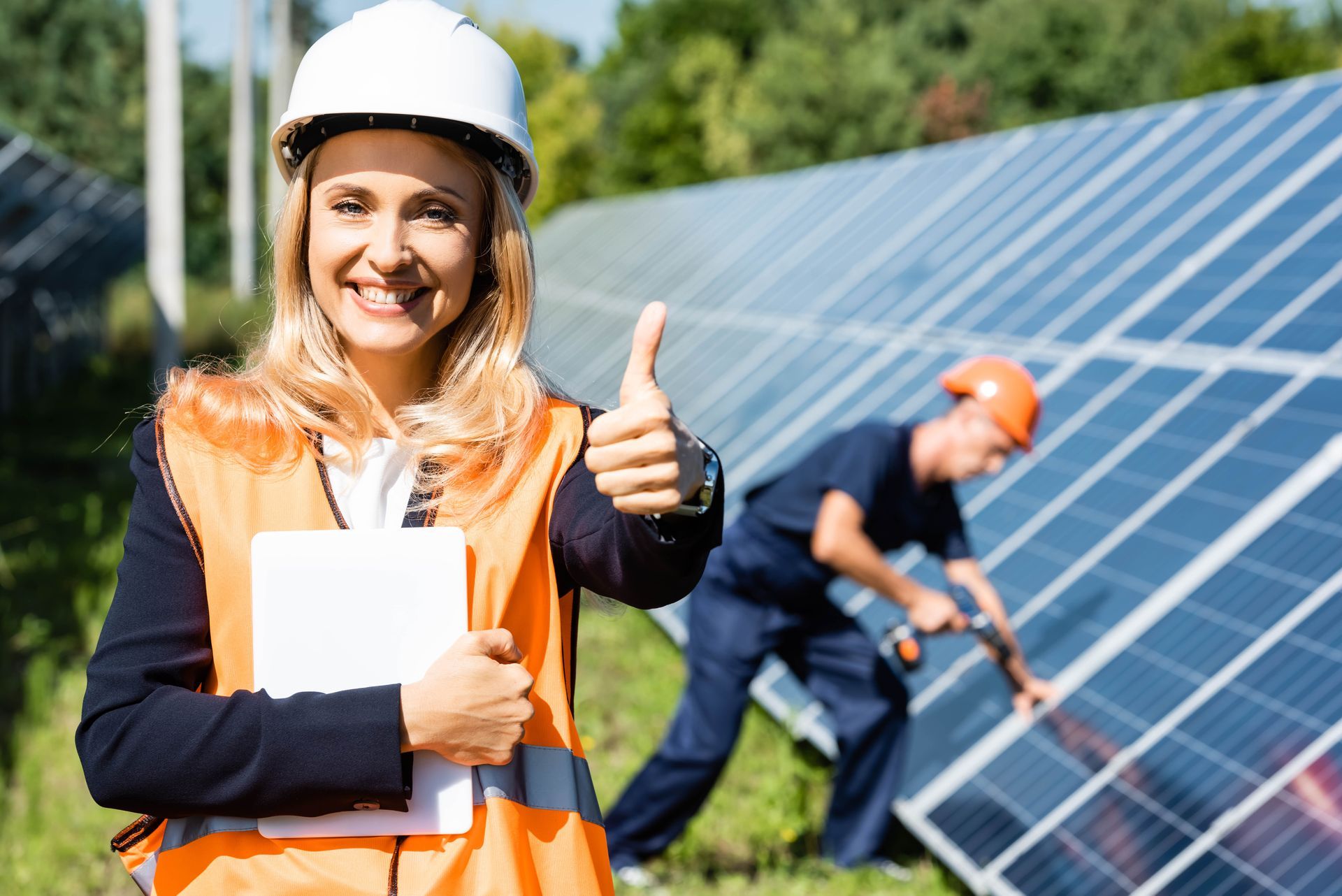 Woman in hard hat gives thumbs up, holding tablet near solar panels with worker in the background.