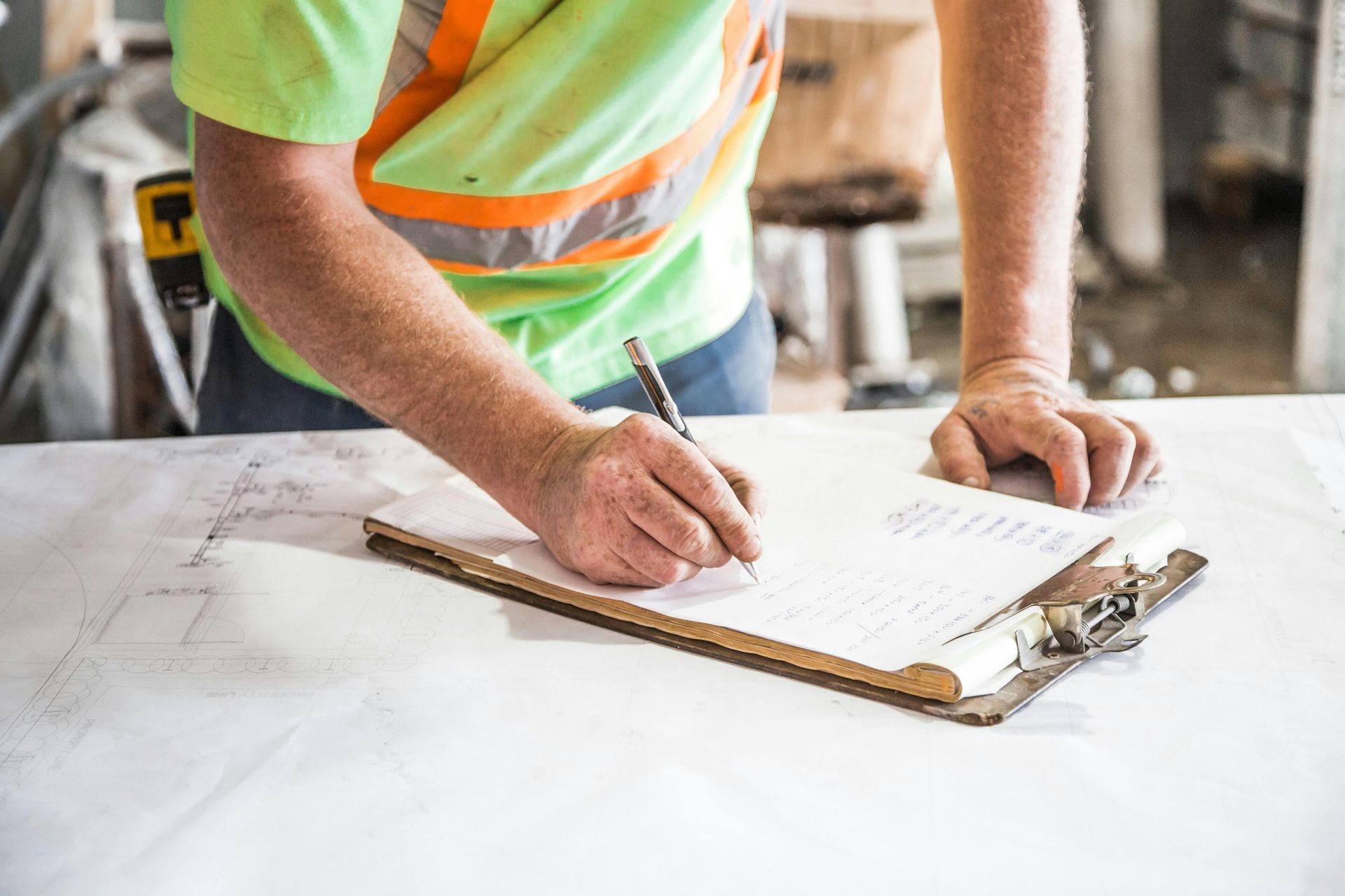 Construction worker in safety vest writing on clipboard, standing at a white surface.