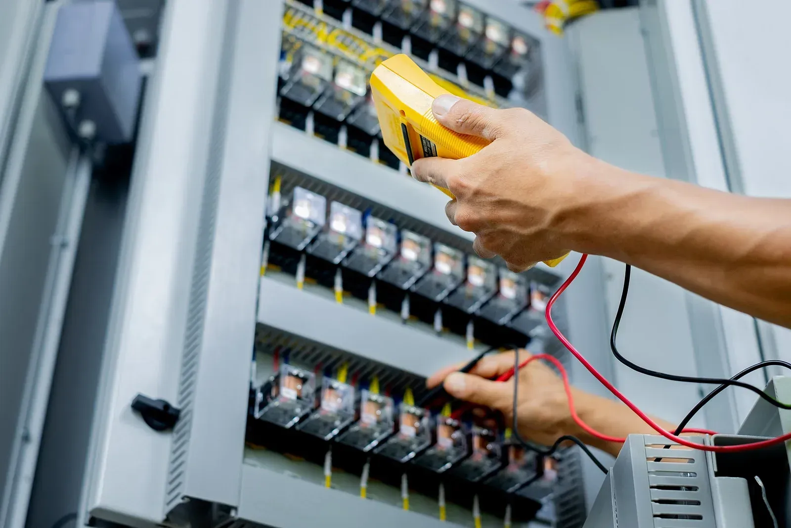 A person using a yellow multimeter to test electrical components in a gray control panel.