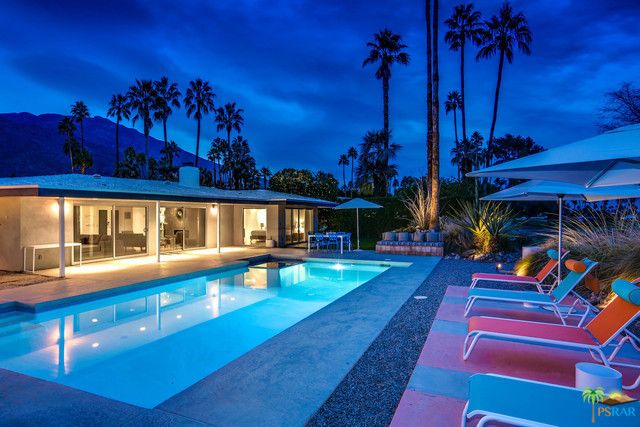 A large swimming pool is surrounded by chairs and umbrellas in front of a house.