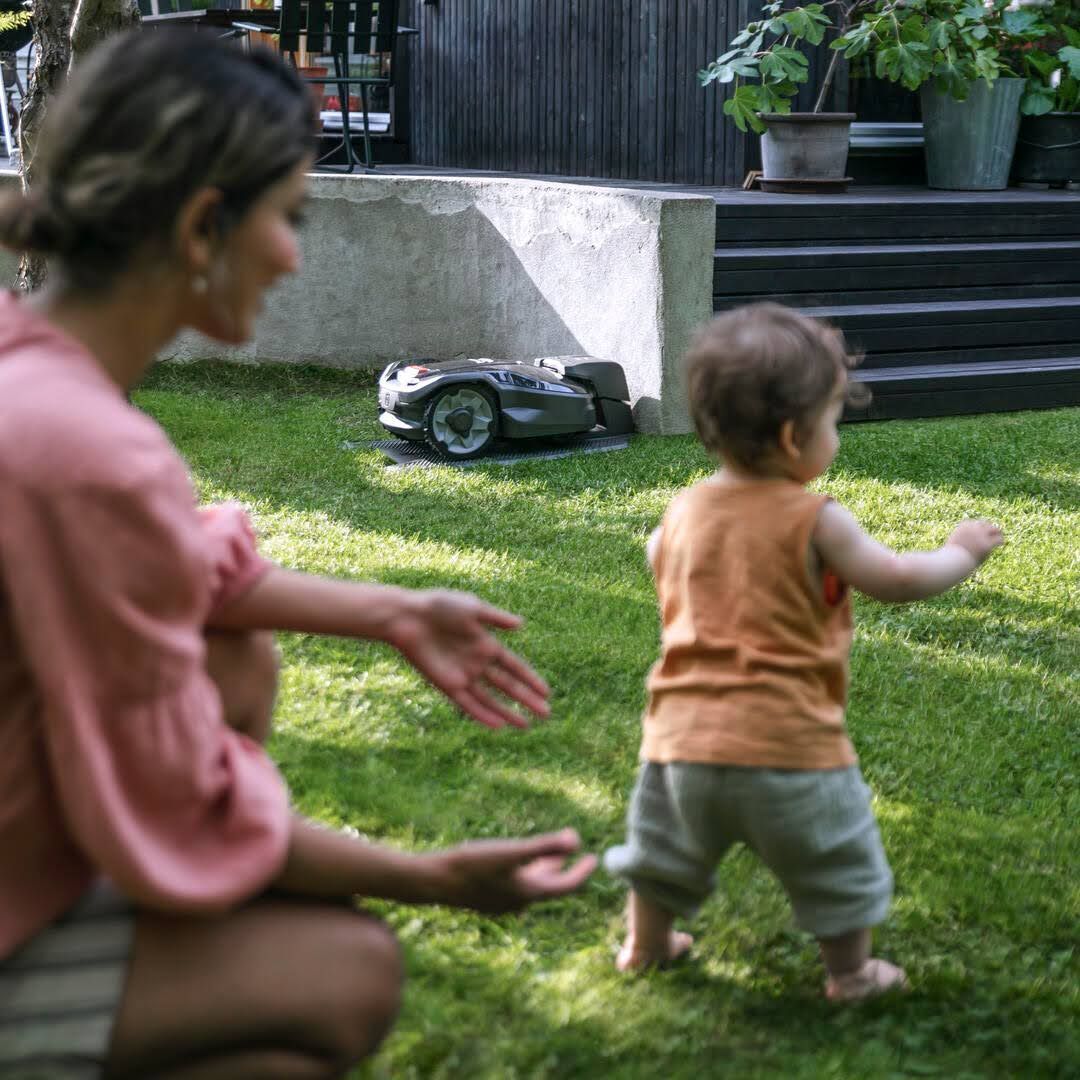 A Woman And A Child Are Playing In The Grass With A Robotic Lawn Mower In The Background — Mid Coast Mowers In Taree, NSW