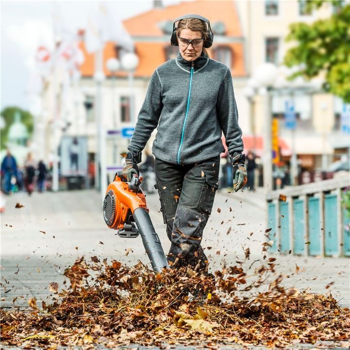 A Woman Is Blowing Leaves On The Sidewalk With A Leaf Blower — Mid Coast Mowers In Taree, NSW