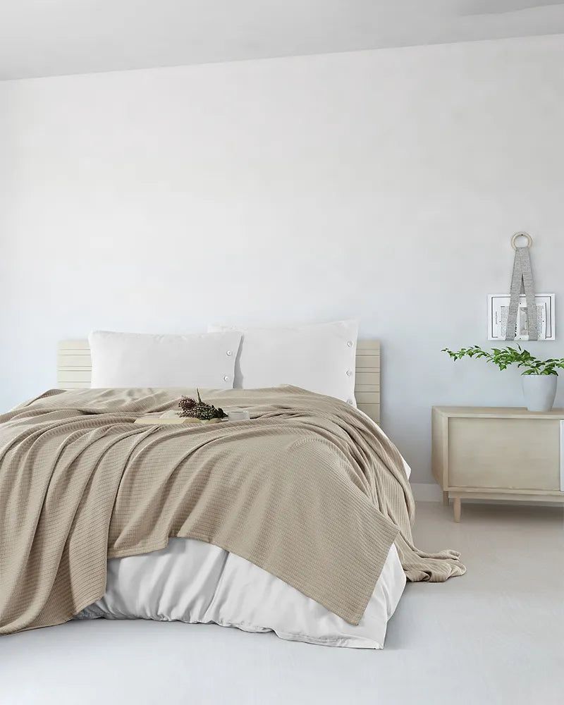 Minimal bedroom with a beige bedspread, white pillows, and a light wood nightstand against white walls.