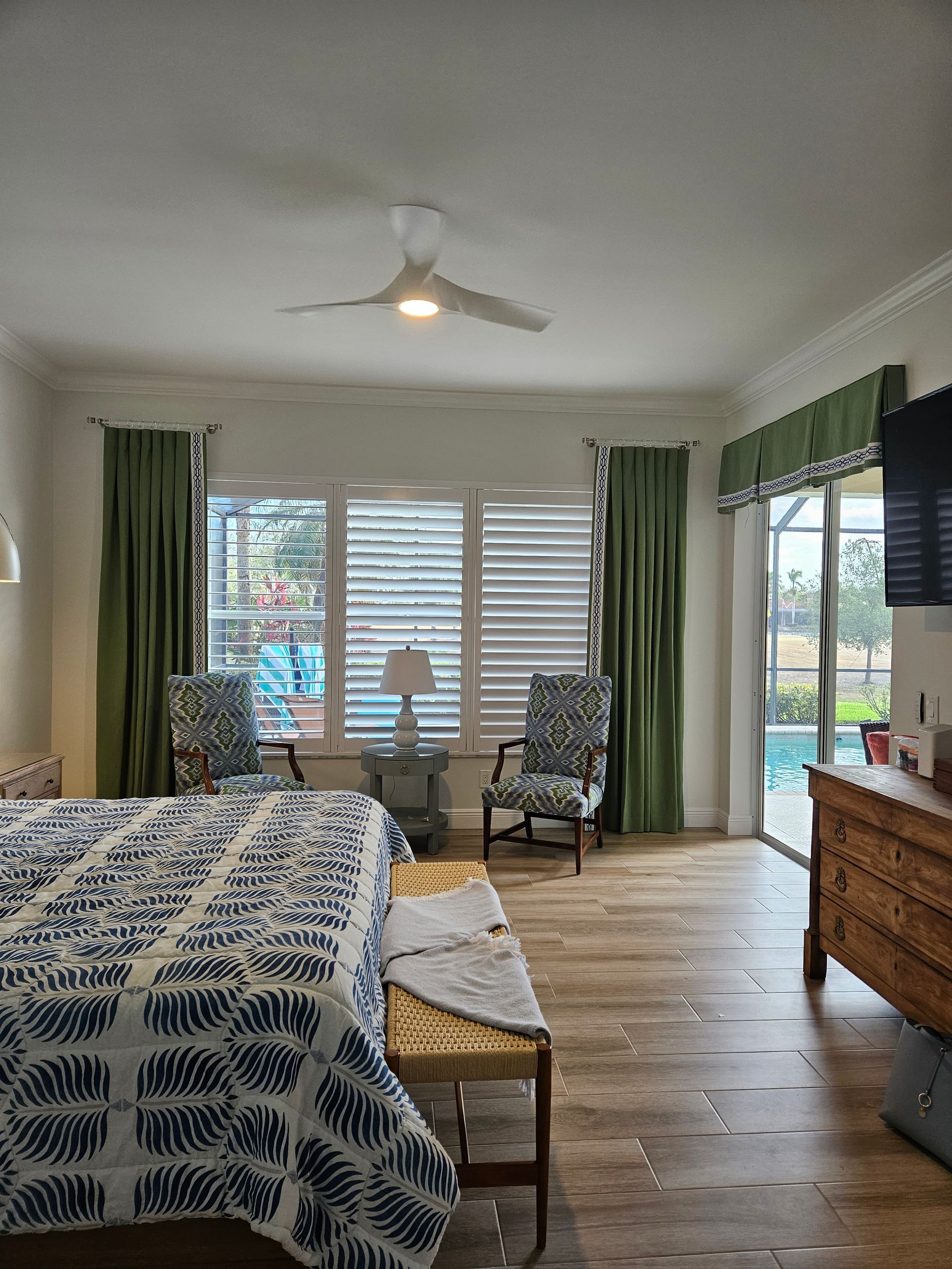 Bright bedroom with bed, wood dresser, green curtains, ceiling fan, and balcony doors.