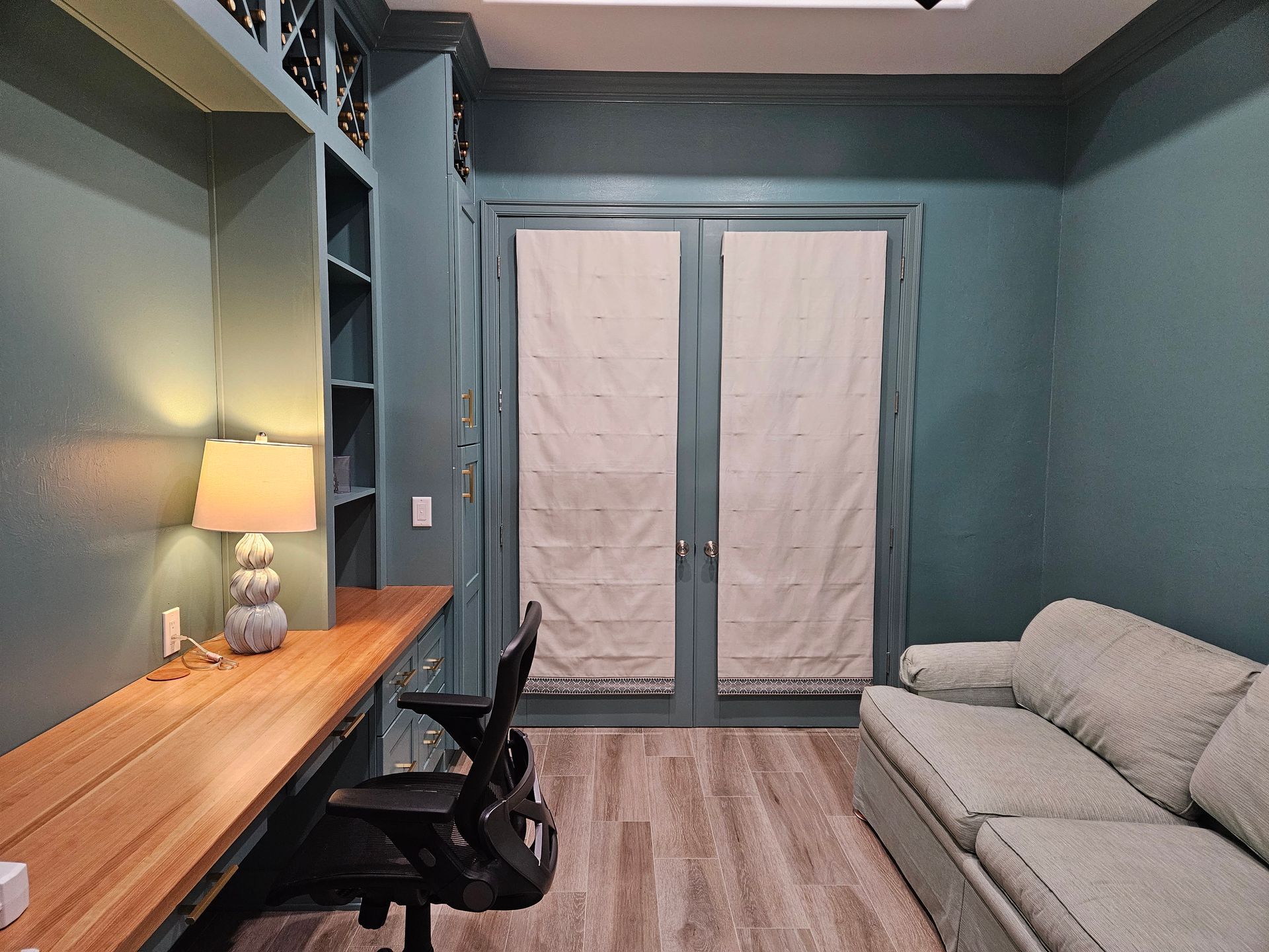 Compact living room with blue walls, desk, lamp, black chair, gray sofa, and French doors covered by curtains
