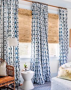 Blue patterned curtains frame a bright bedroom with woven shades, a white pedestal table, and a wicker chair.