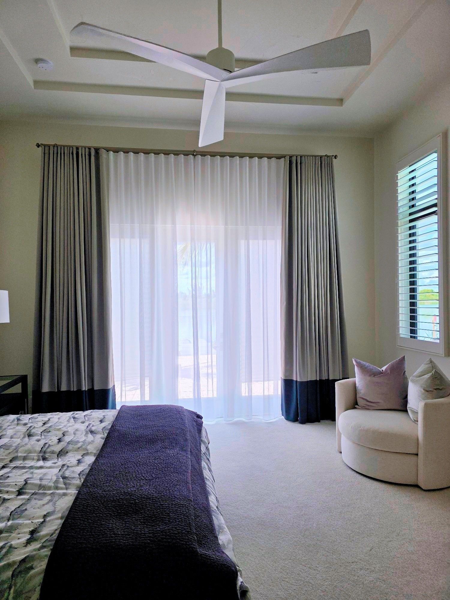 Bright bedroom with a ceiling fan, sheer curtains, carpeted floor, and a beige chair by the window.