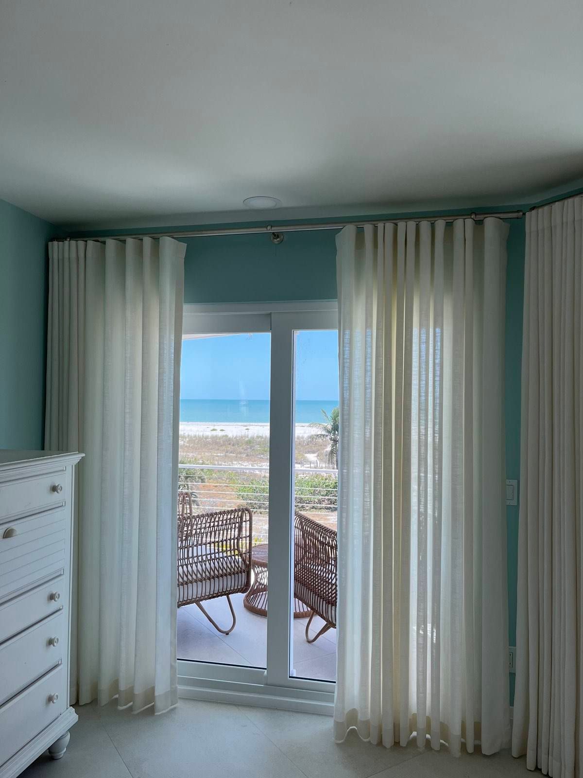Bright bedroom with white curtains framing ocean-view glass doors and a balcony with chairs