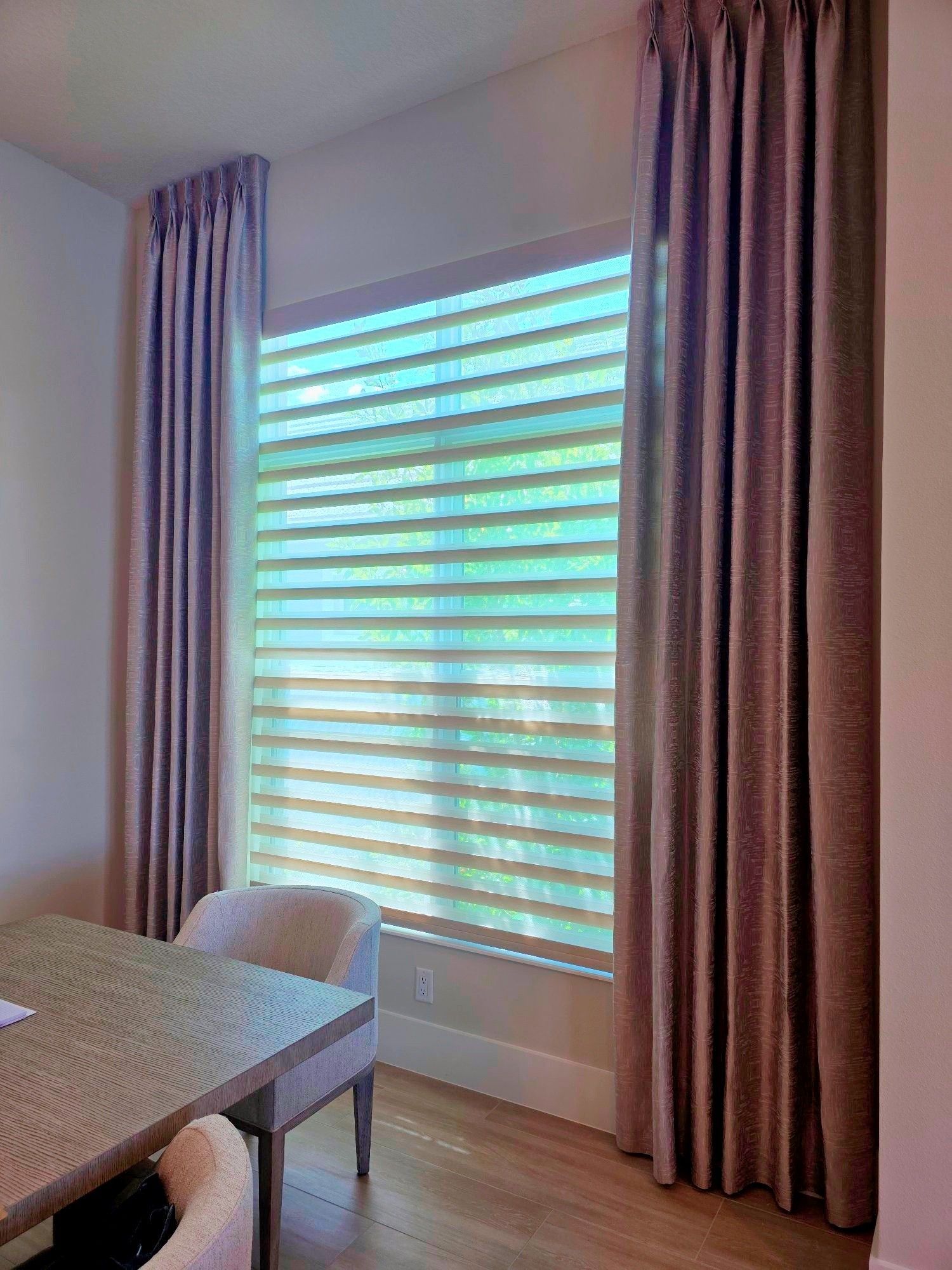 Dining room with a table, upholstered chair, and tall window with sheer curtains and blinds.
