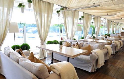 Bright beachside lounge with white sofas, curtains, and tables overlooking the water