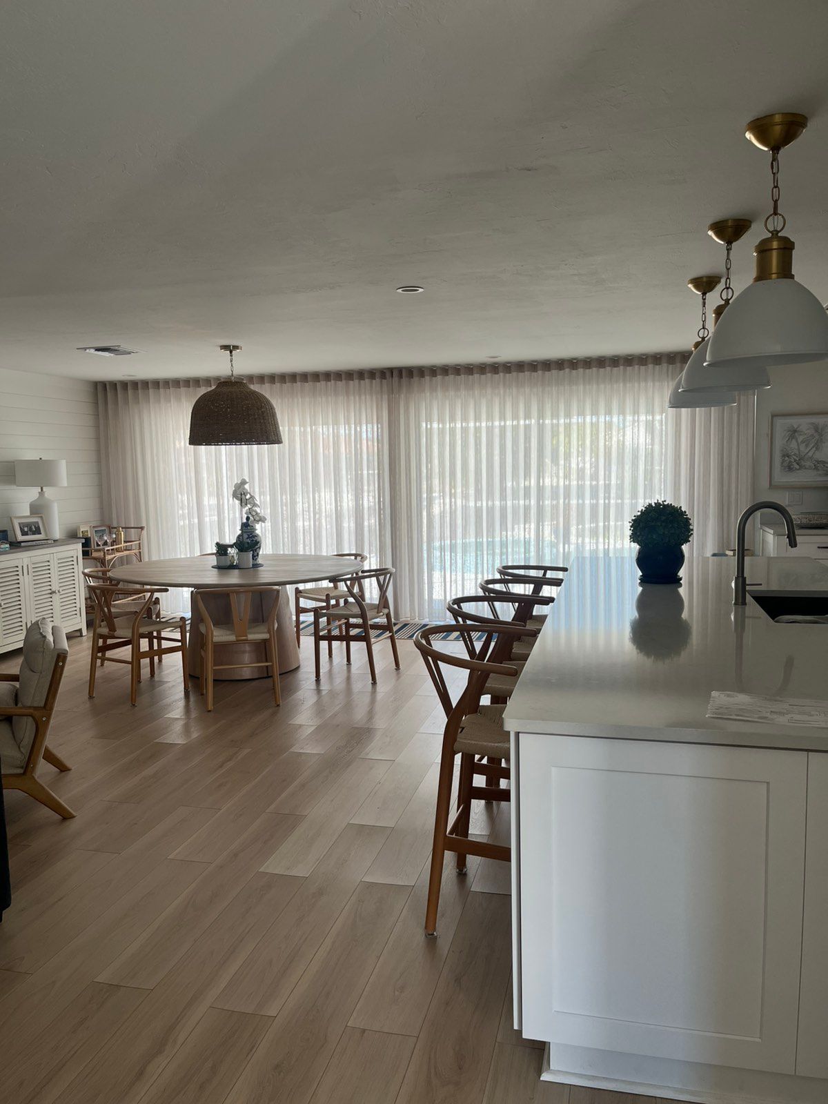 Bright open kitchen and dining area with wood floors, white island, and large windows with sheer curtains.