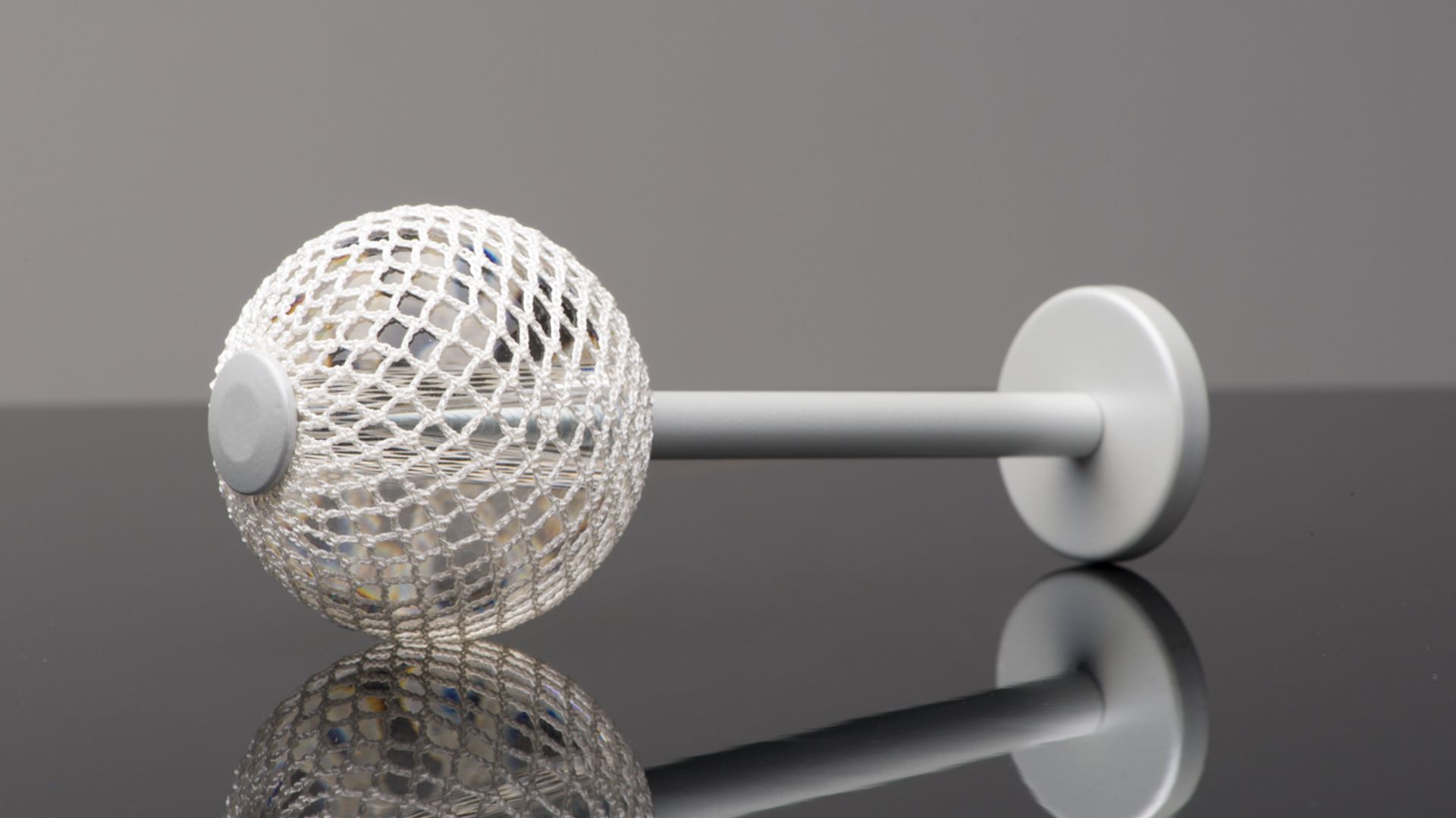 White mesh ball connected to a gray rod with circular end caps on a reflective surface