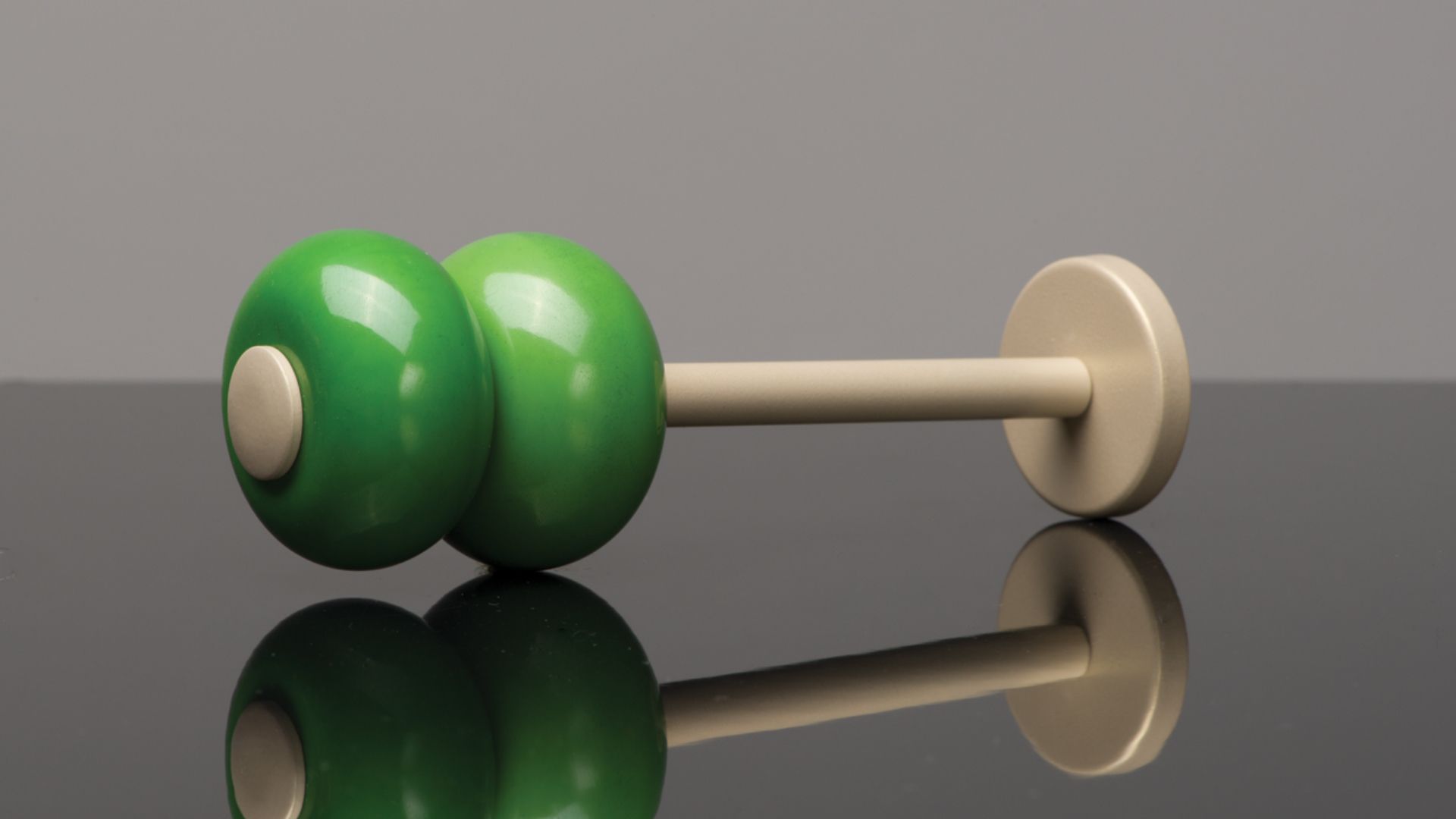 Green dumbbell with beige handle and ends, resting on a glossy reflective surface against a gray background
