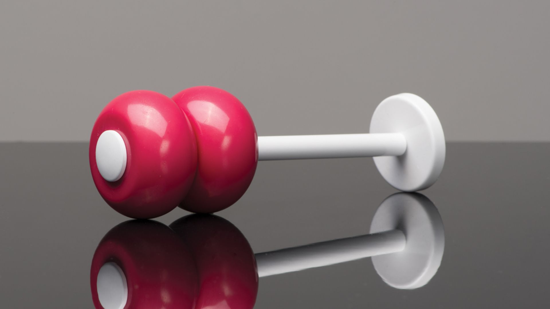 Red dumbbell with white handle and end caps on a glossy black surface, with a reflection.