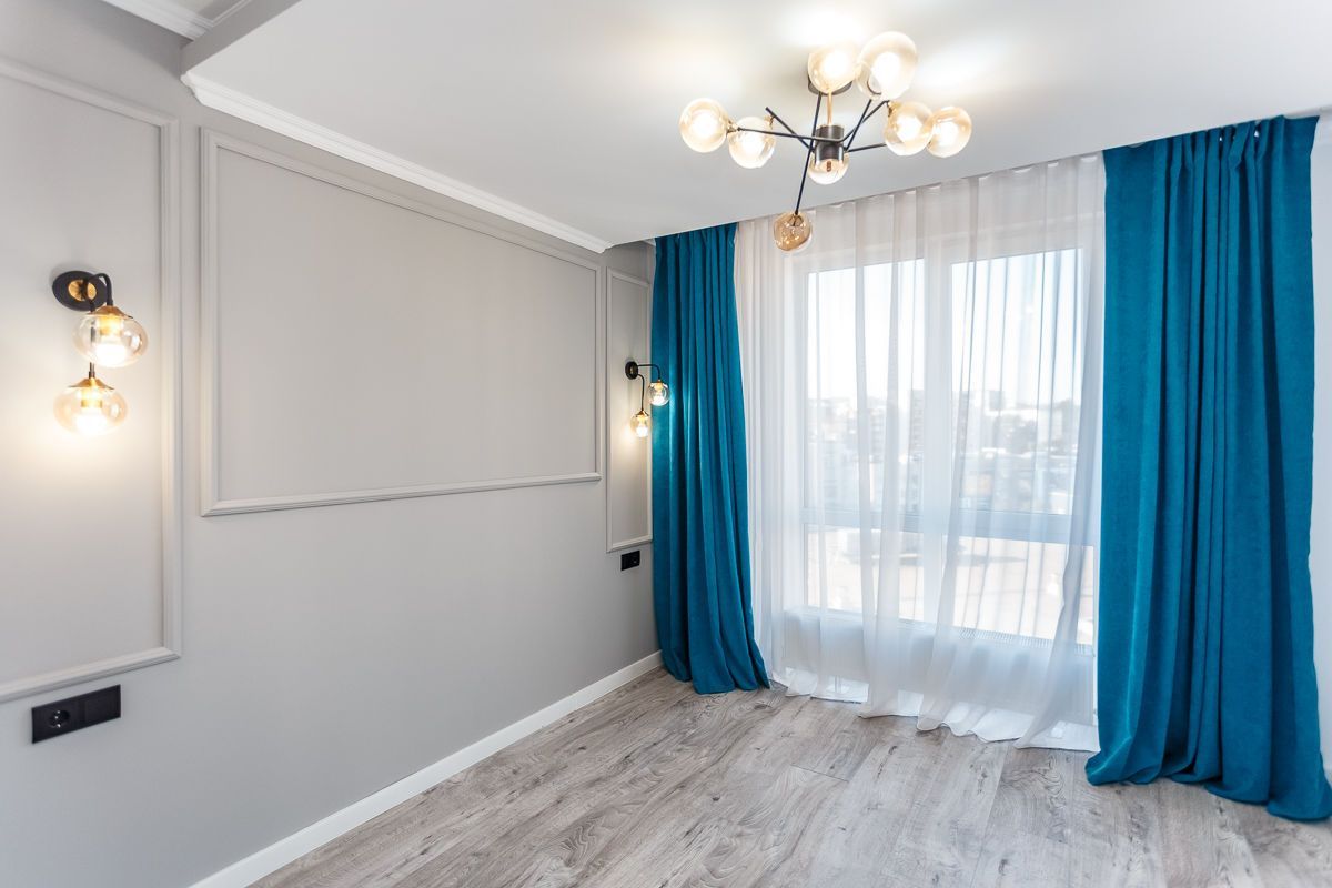 Bright empty room with gray walls, white curtains, blue drapes, and a chandelier near a window.