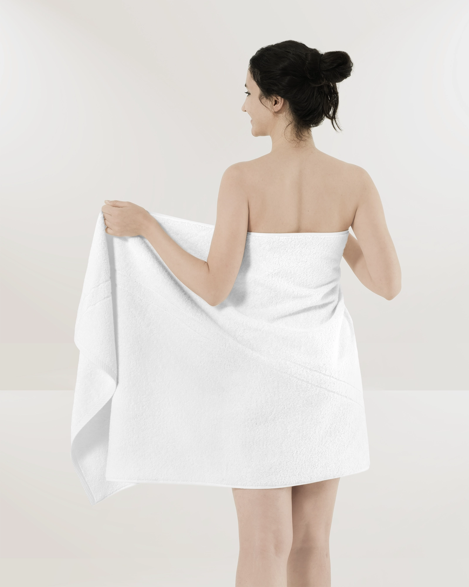 Woman in a white strapless towel, viewed from behind, holding a white towel against a plain background.