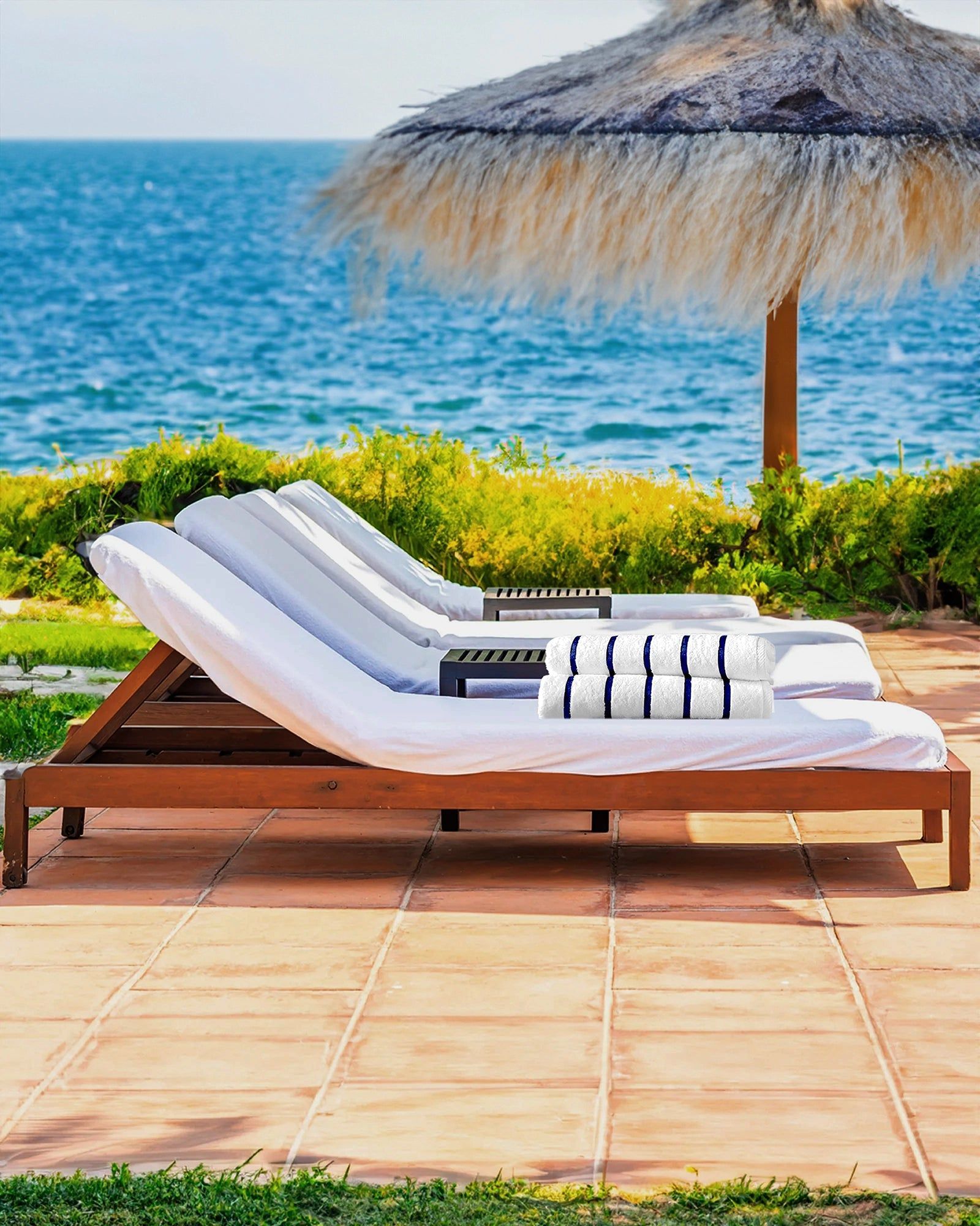 Lounge chair with white cushion and striped towel on a seaside patio beside a thatched umbrella and blue ocean.