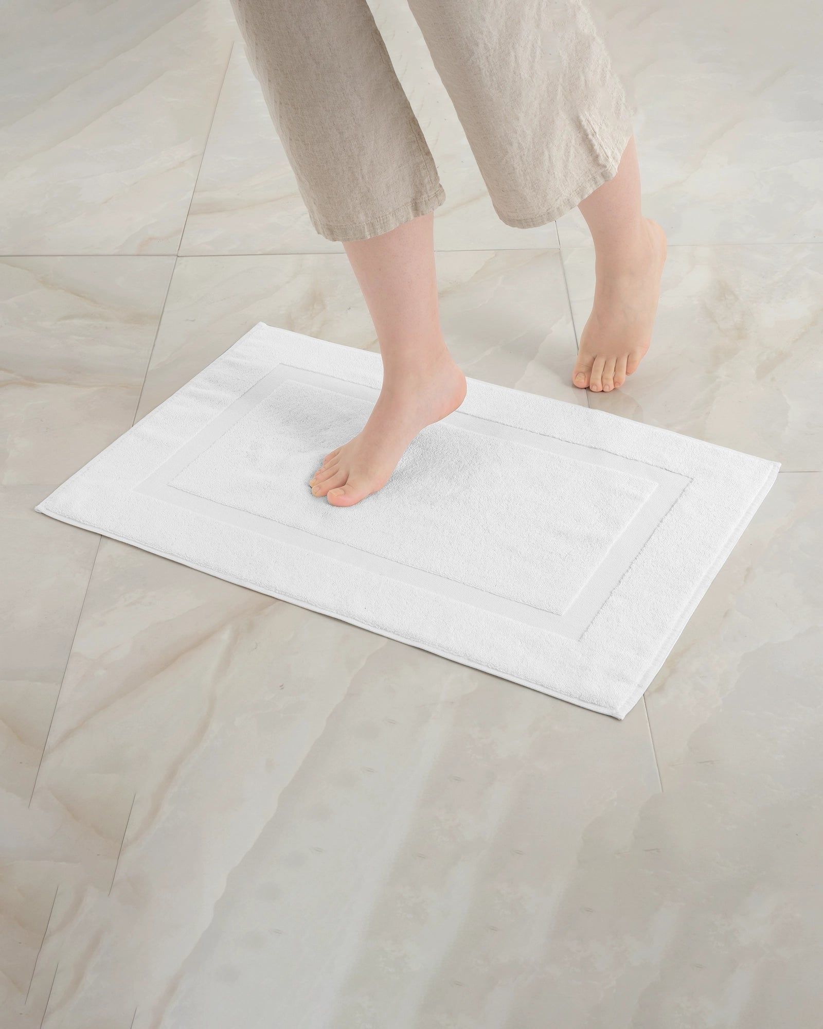 Bare feet stepping on a white bath mat on a light tiled floor.