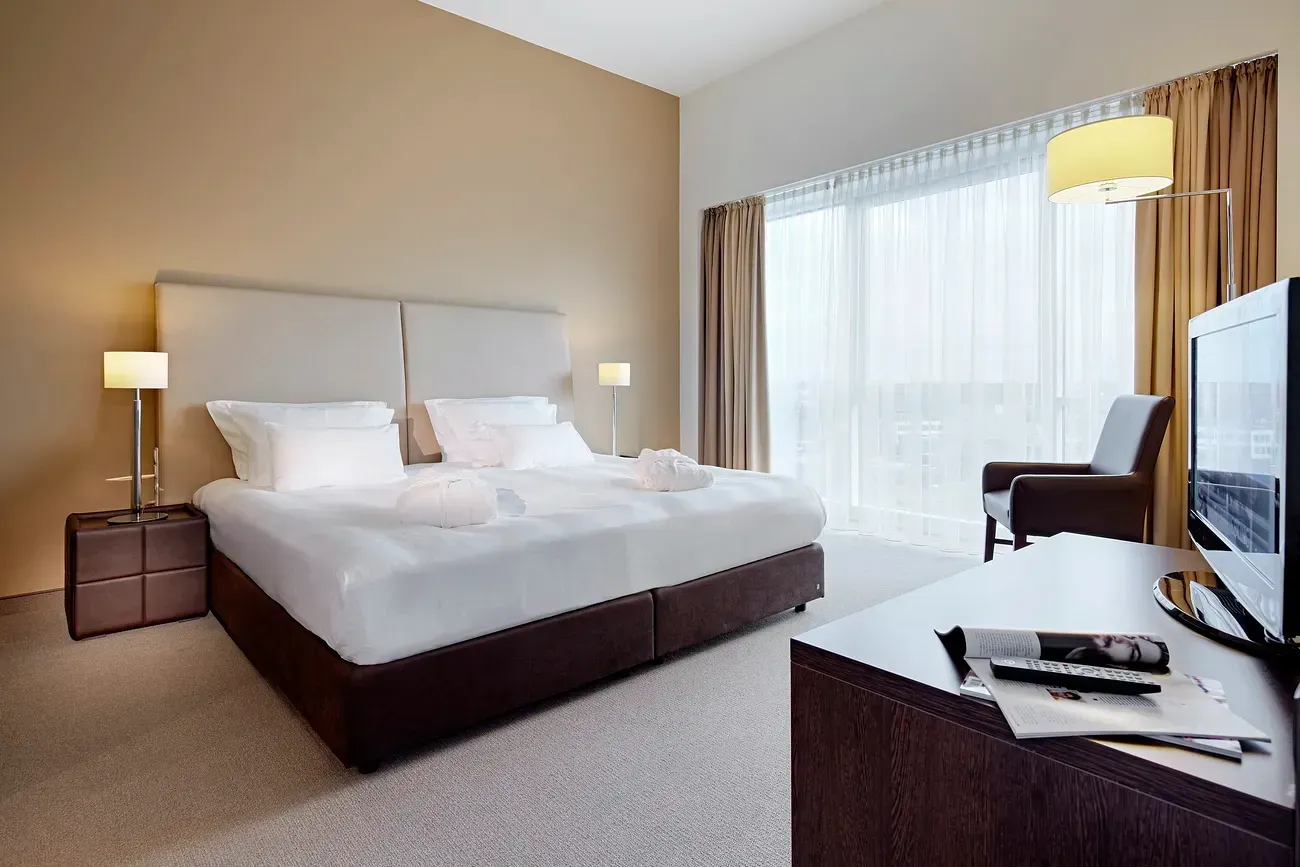 Modern hotel bedroom with king bed, bedside lamps, desk, chair, TV, and large window with sheer curtains