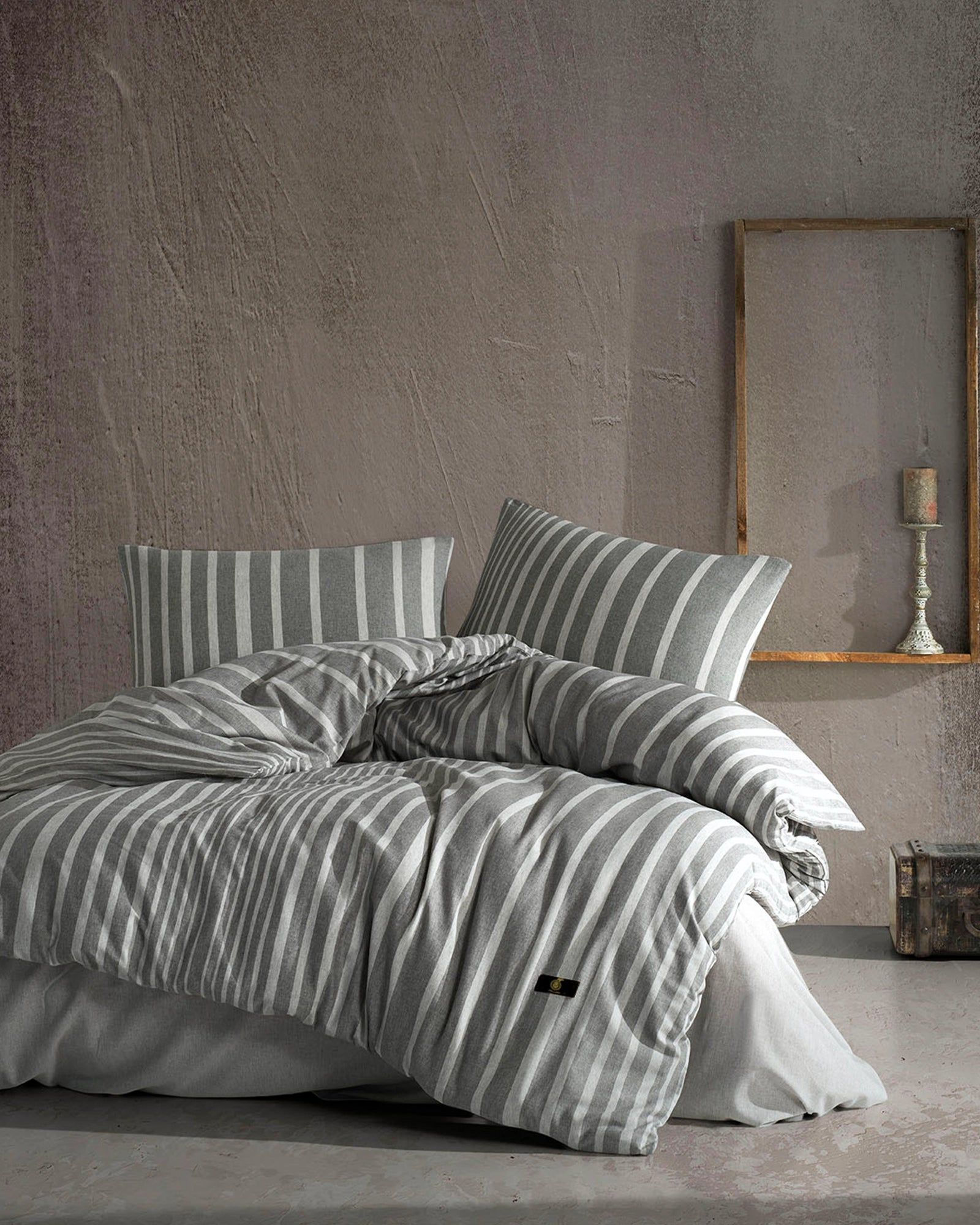 Gray striped bedding on a neatly made bed in a minimalist bedroom with a wooden mirror and lamp.