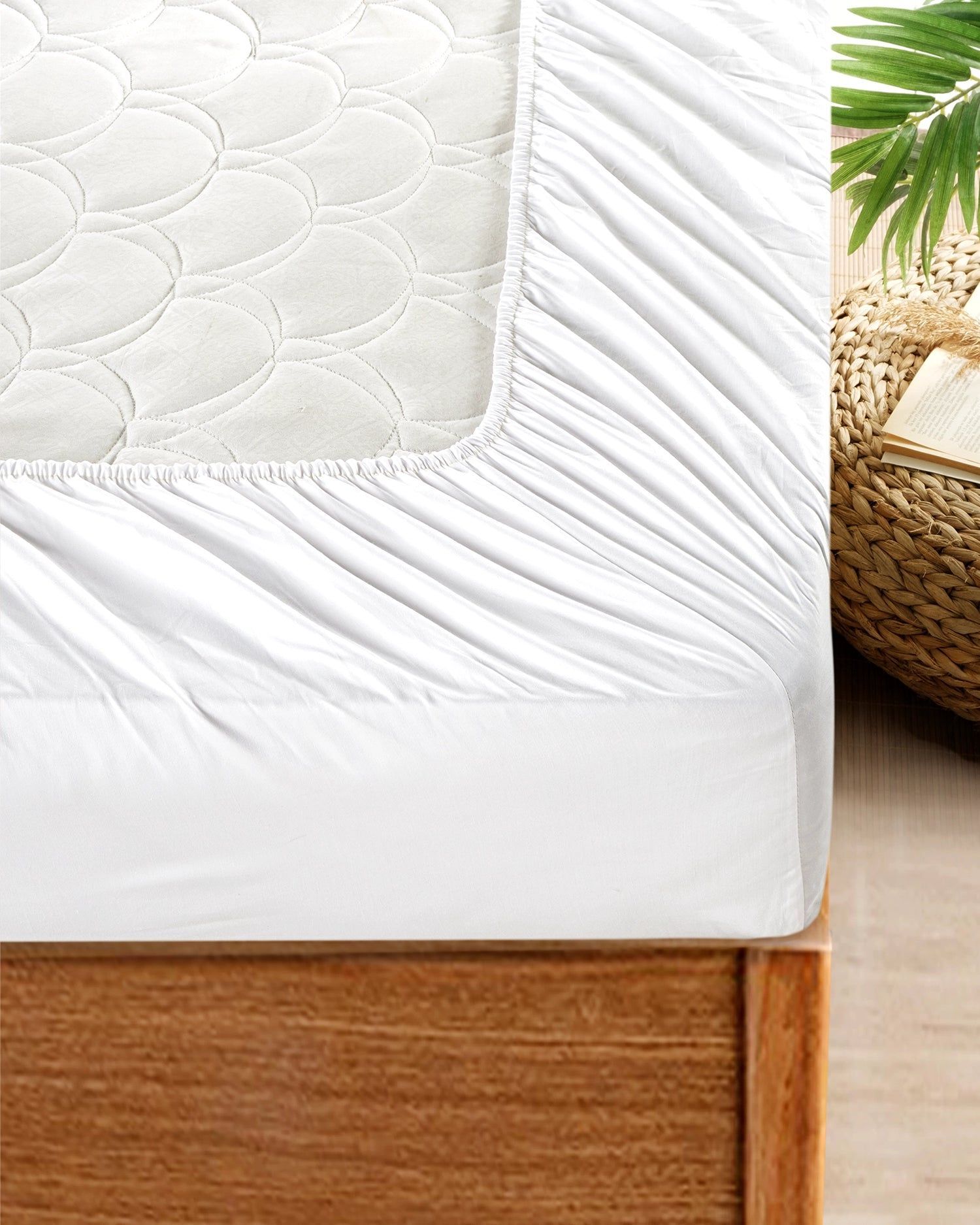 Close-up of a white mattress on a wooden bed frame beside a wicker basket and palm leaves.